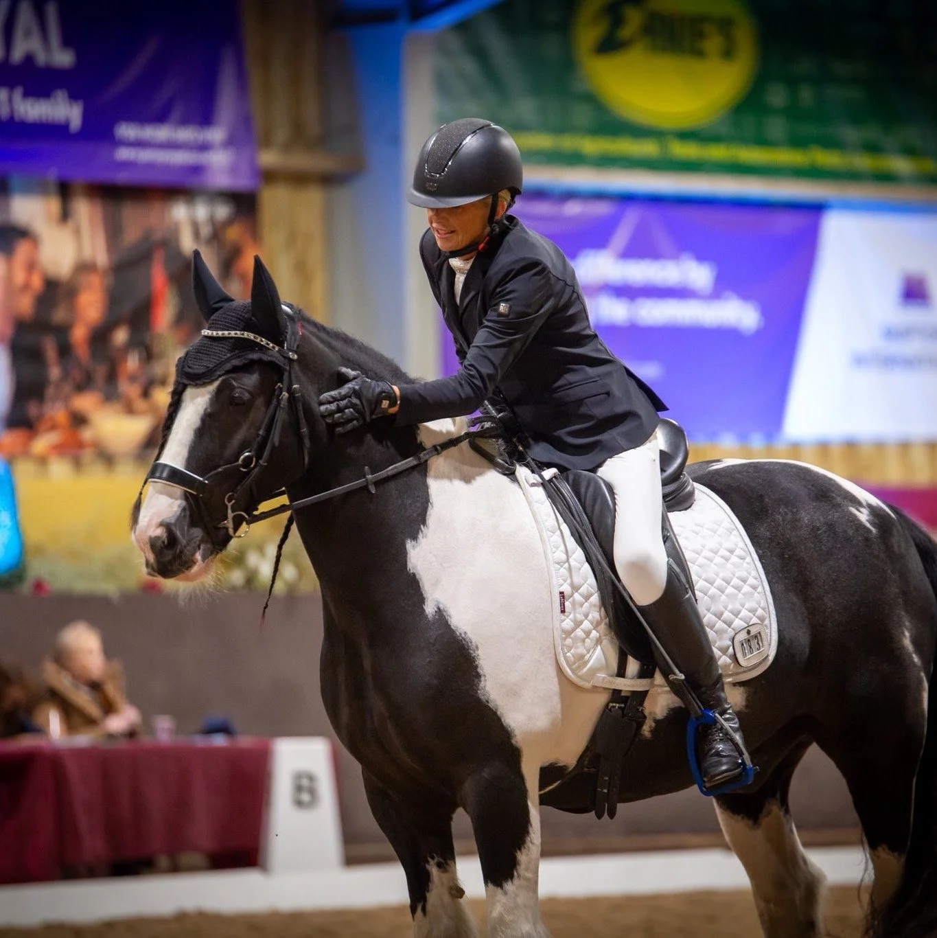 A woman dressed in equestrian riding attire, including a helmet, jacket, and gloves, is smiling and patting the neck of a black and white pinto horse inside an indoor riding arena.