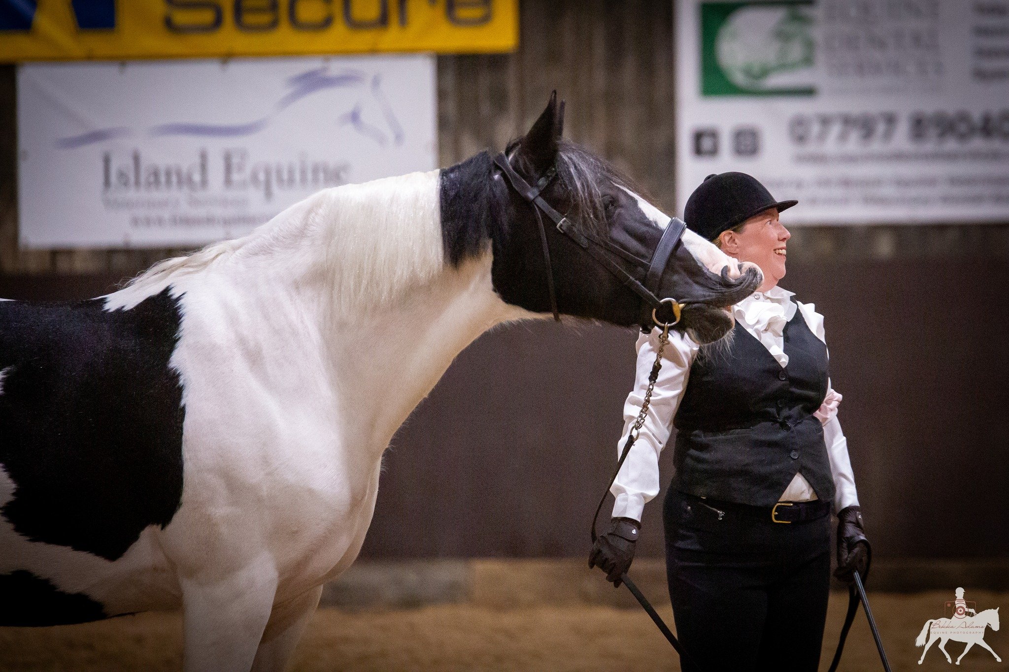 A woman dressed in equestrian attire holding the reins of a black and white horse inside an indoor riding arena.