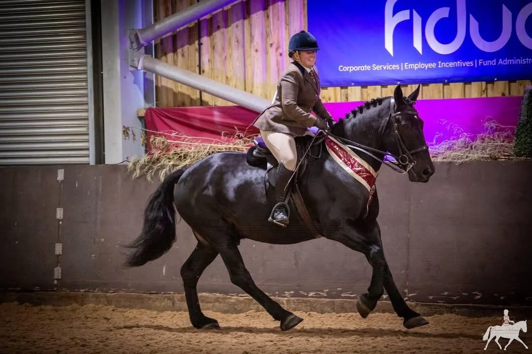 A woman in equestrian attire riding a black horse inside an indoor riding arena.