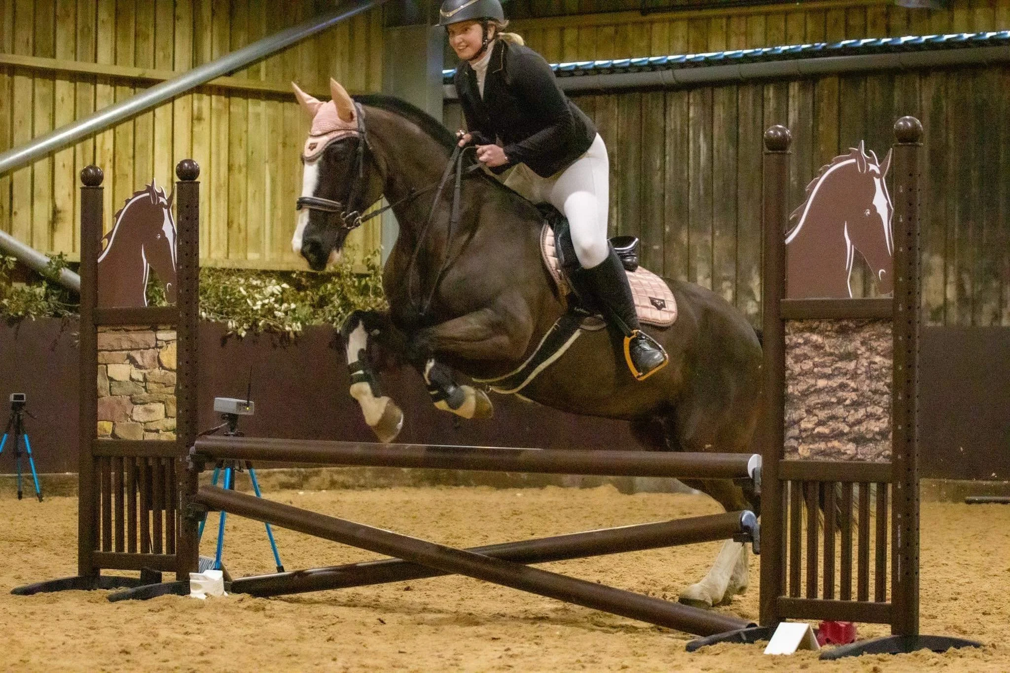 A woman riding a dark brown horse while jumping over an obstacle in an indoor equestrian arena.
