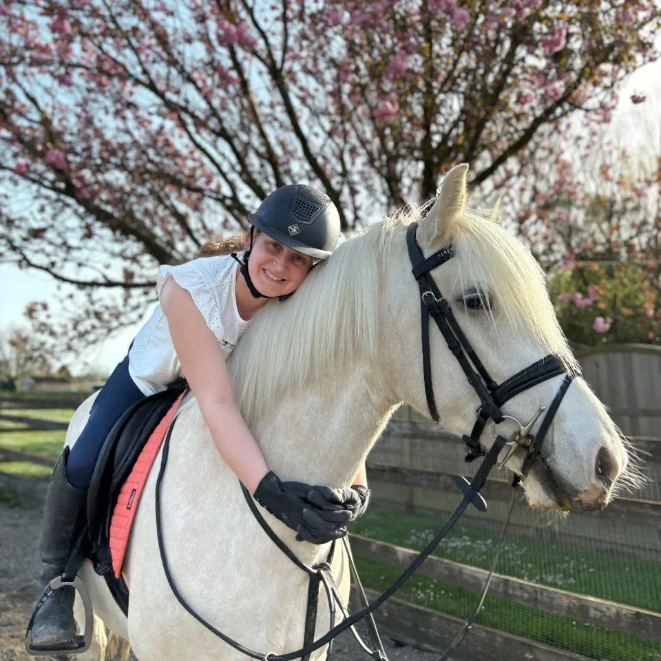 A young girl in equestrian gear riding a white horse, smiling, with cherry blossom trees in bloom in the background.
