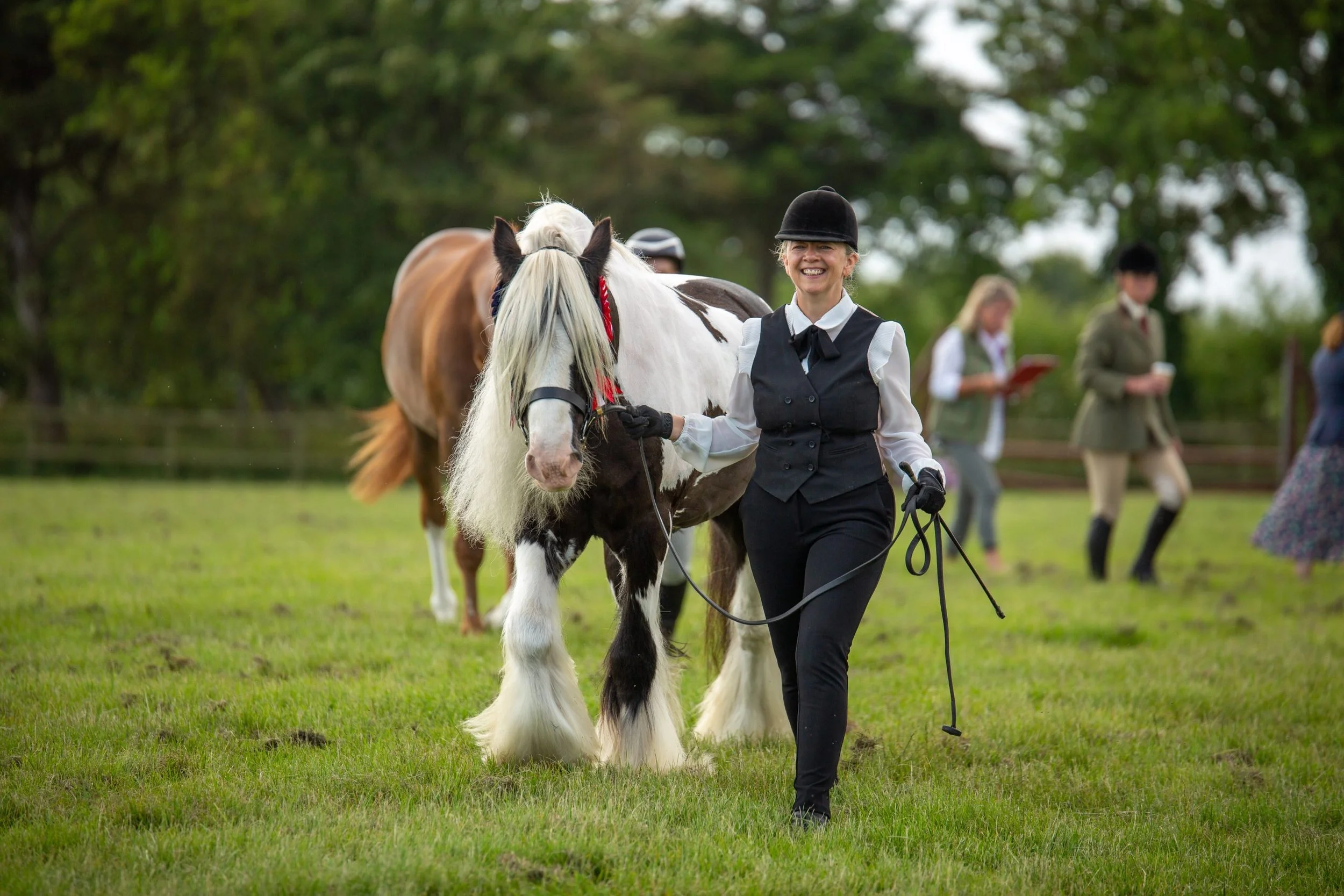 A woman in formal riding attire walking a black and white pinto horse on a grassy field. In the background, other women are standing and reading papers, surrounded by green trees.