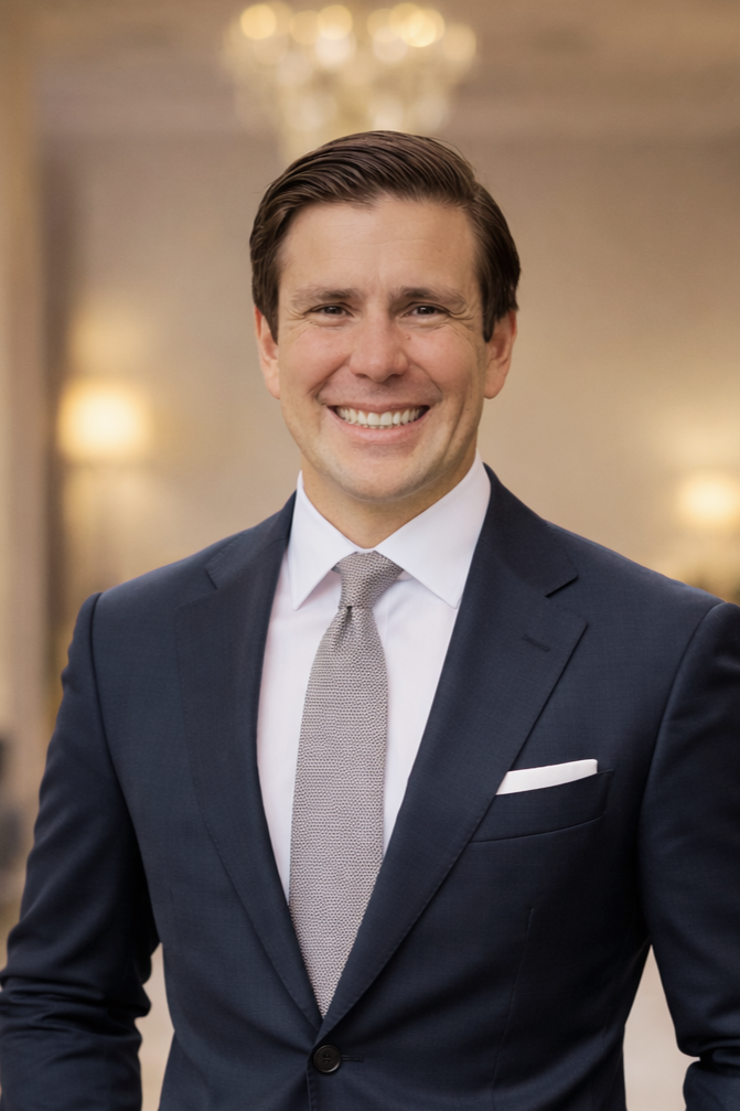 A smiling man in a dark suit, white shirt, and gray tie, standing indoors with warm lighting and blurred background.