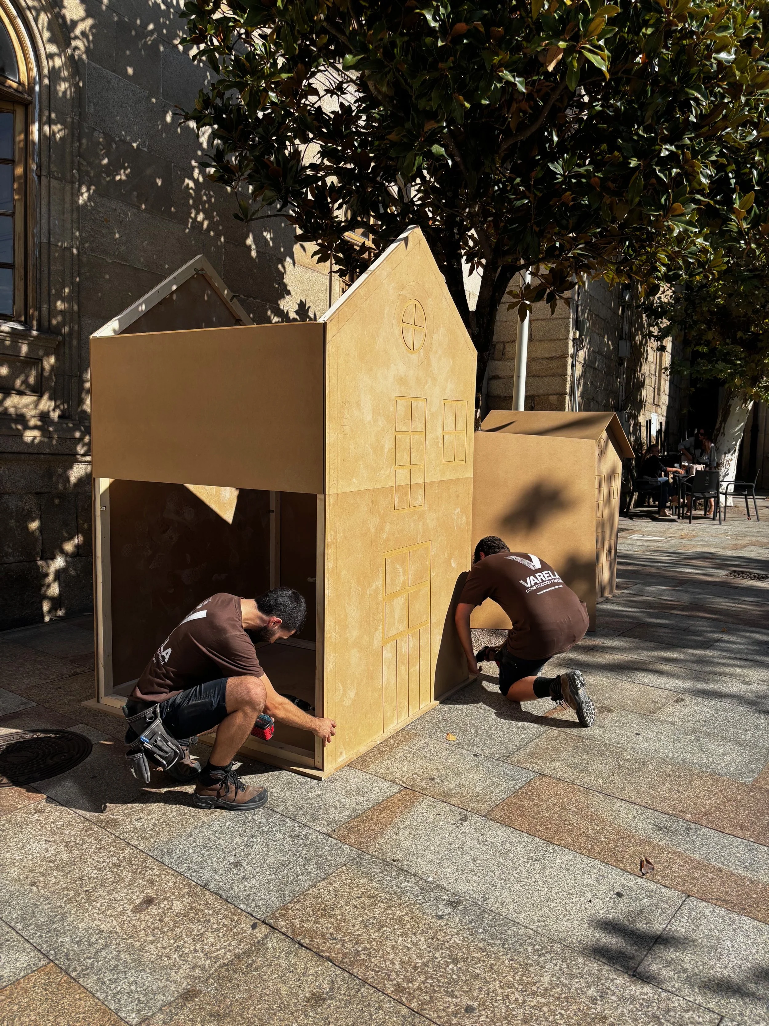 Dos personas armando una pequeña casita de madera en la calle, con un árbol y algunas personas en el fondo, en un día soleado.