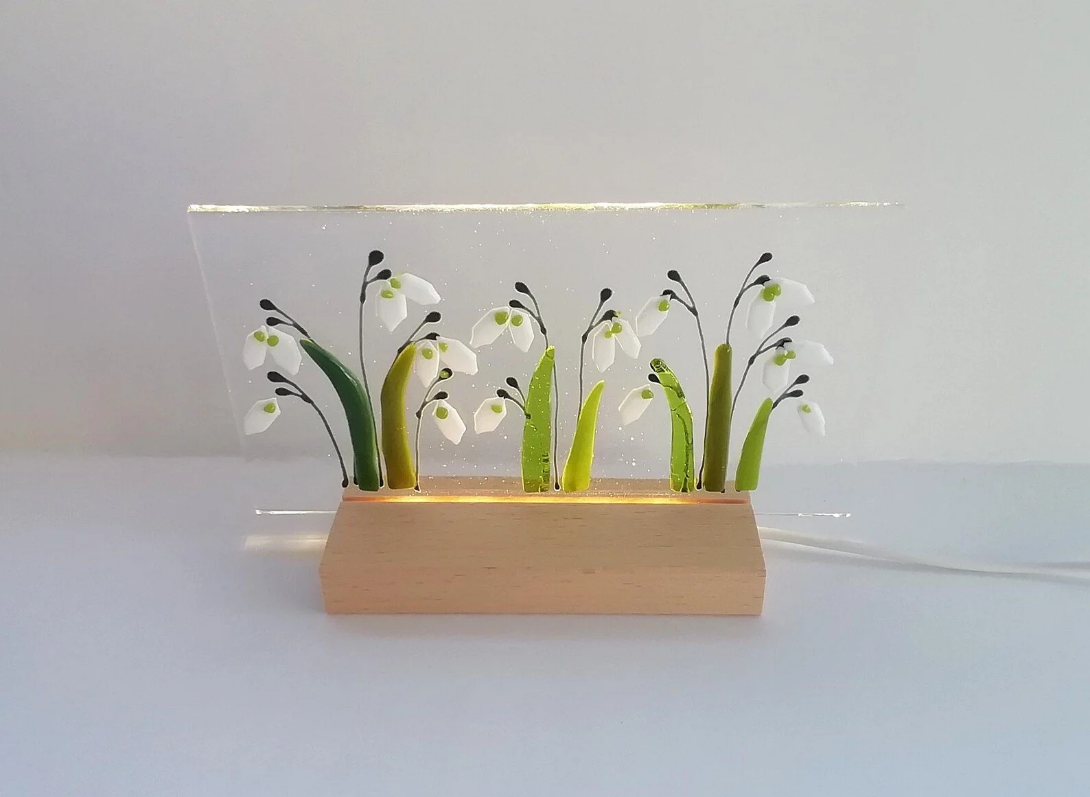 Decorative lighted wood base with small artificial snowdrop flowers arranged in a row behind a clear acrylic panel.
