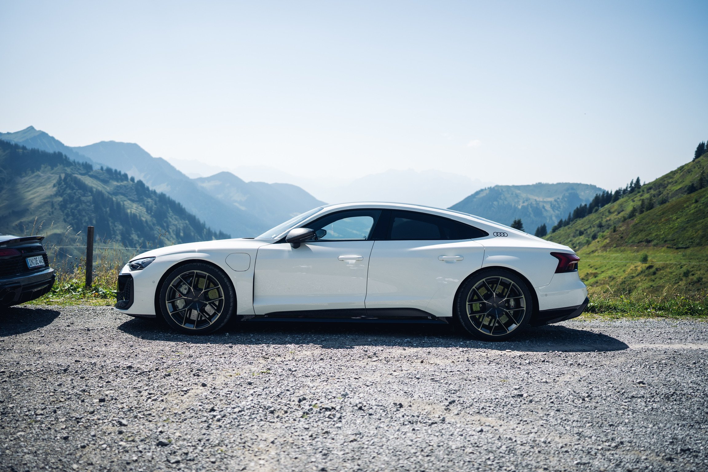Weiße Audi Sportlimousine auf einer Bergstraße im Grünen, Blick auf Berge im Hintergrund, klare Himmel.