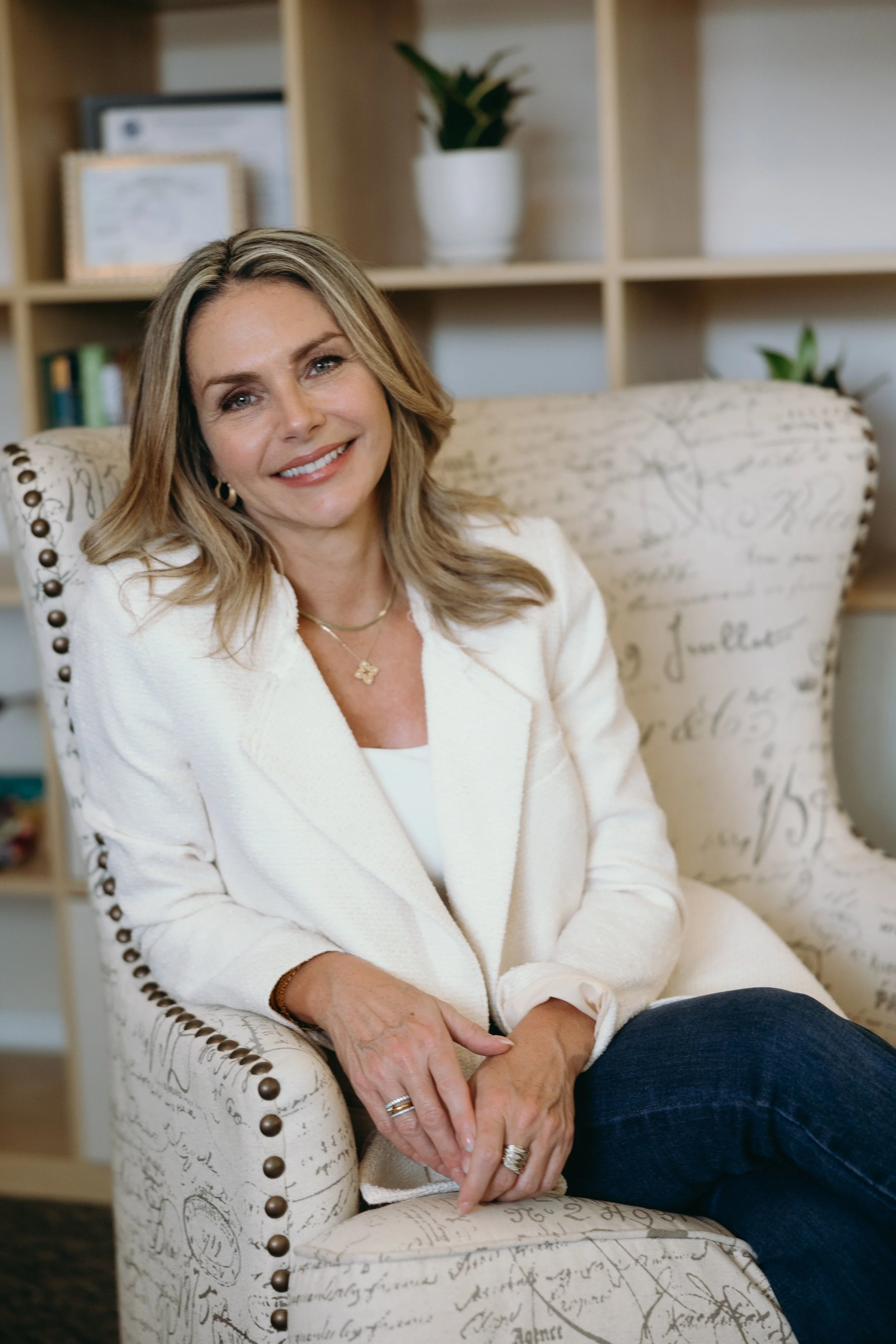 A woman smiling, sitting on a patterned armchair in a room with wooden shelves and plants.