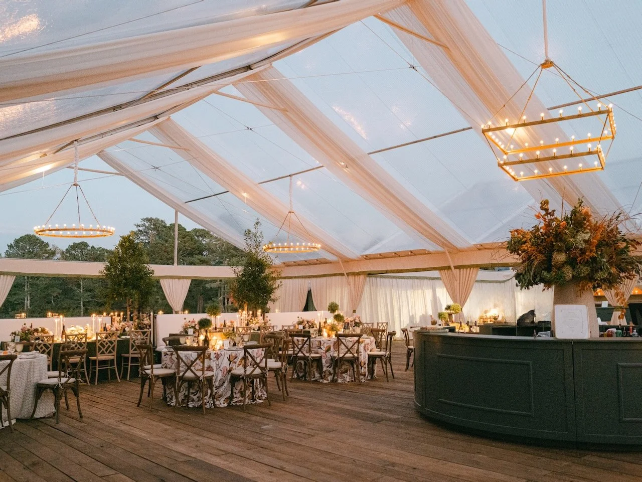 A memorable reception space is created at a family home using a clear tent, soft fabrics, modern chandeliers, circular bar with large floral centerpiece, and exceptionally designed tablescapes for dining
