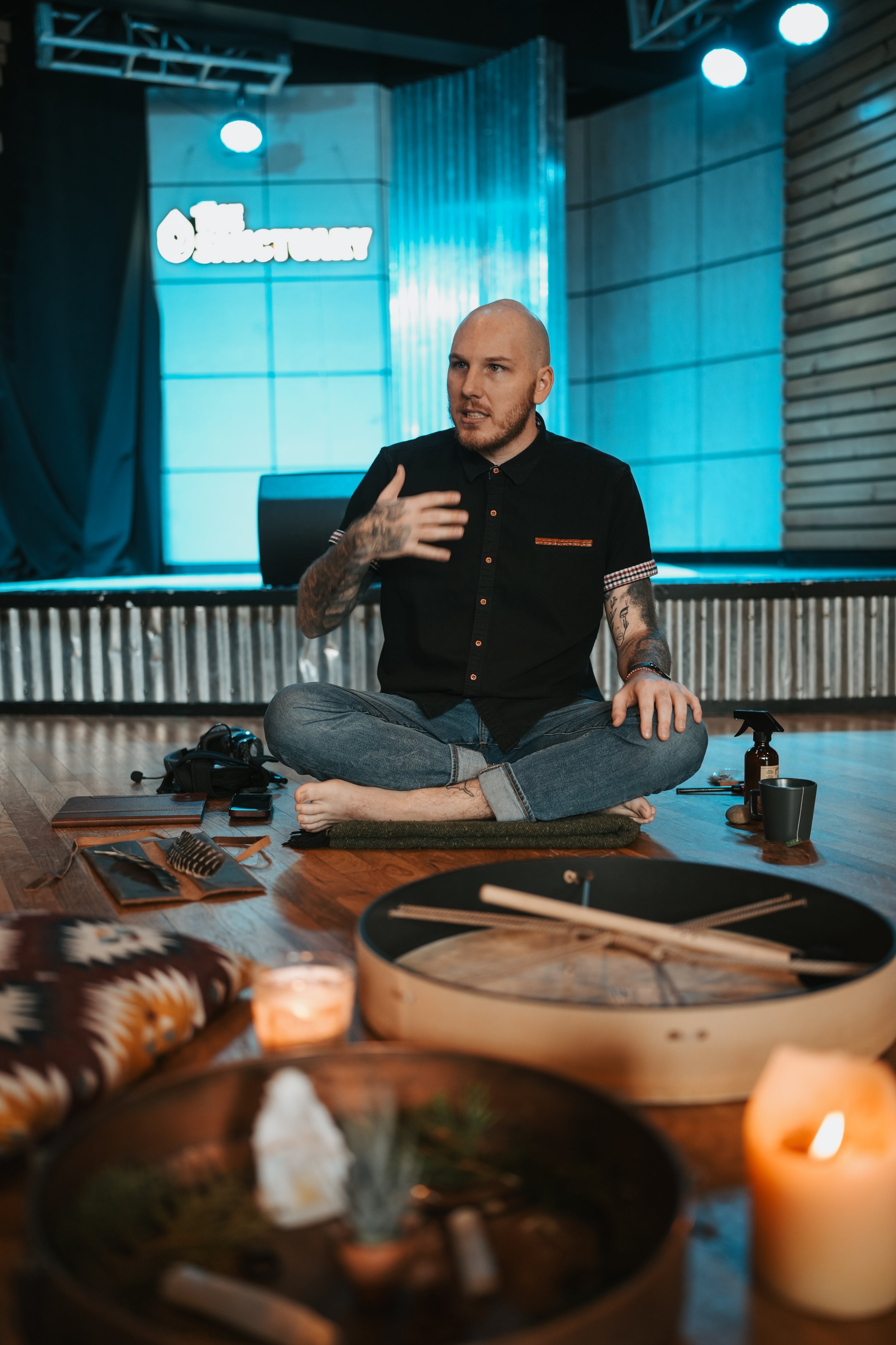 A man with tattoos and a shaved head sitting cross-legged on the floor, speaking and gesturing with his hand. The setting appears to be indoors with a dim, modern ambiance, decorated with candles, a small drum, and musical instruments nearby. There is a stage and a blue-lit backdrop with a sign that reads 'The Outlaw Lounge'.