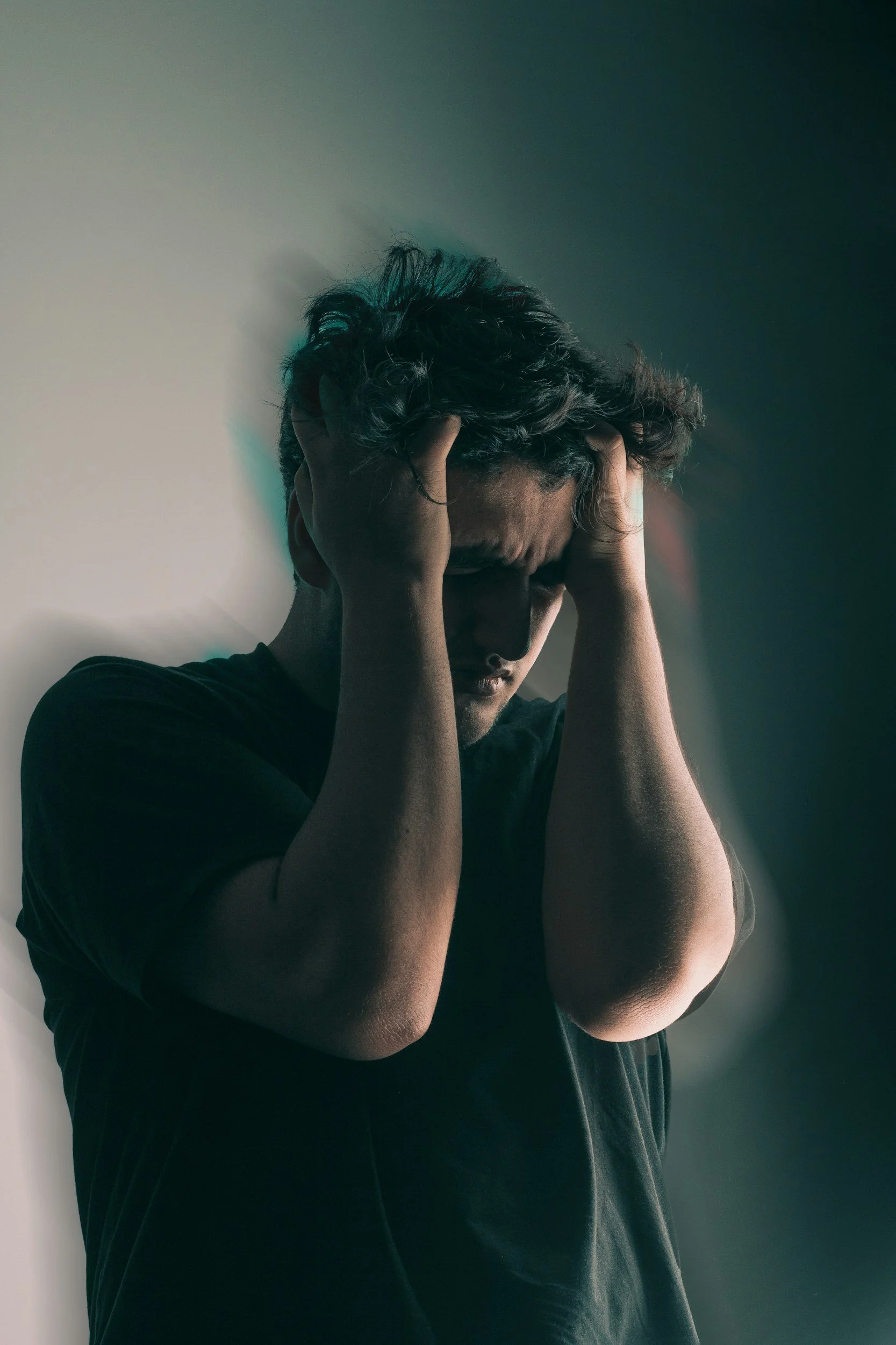 A man with messy hair holds his head with both hands, appearing stressed or distressed, against a plain wall with shadows.