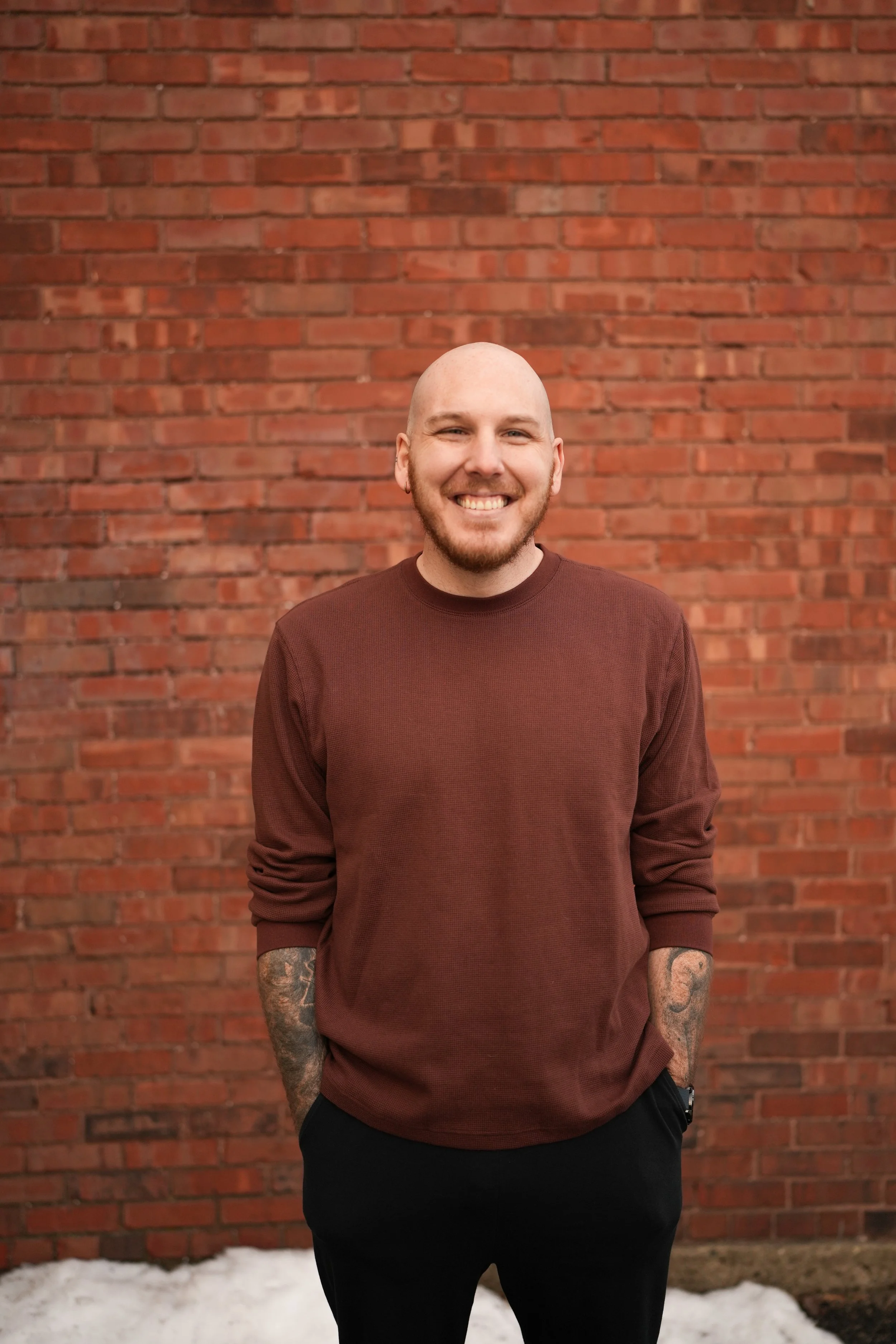 A smiling man with a beard, bald head, and tattoos on his arms, standing outdoors against a brick wall.