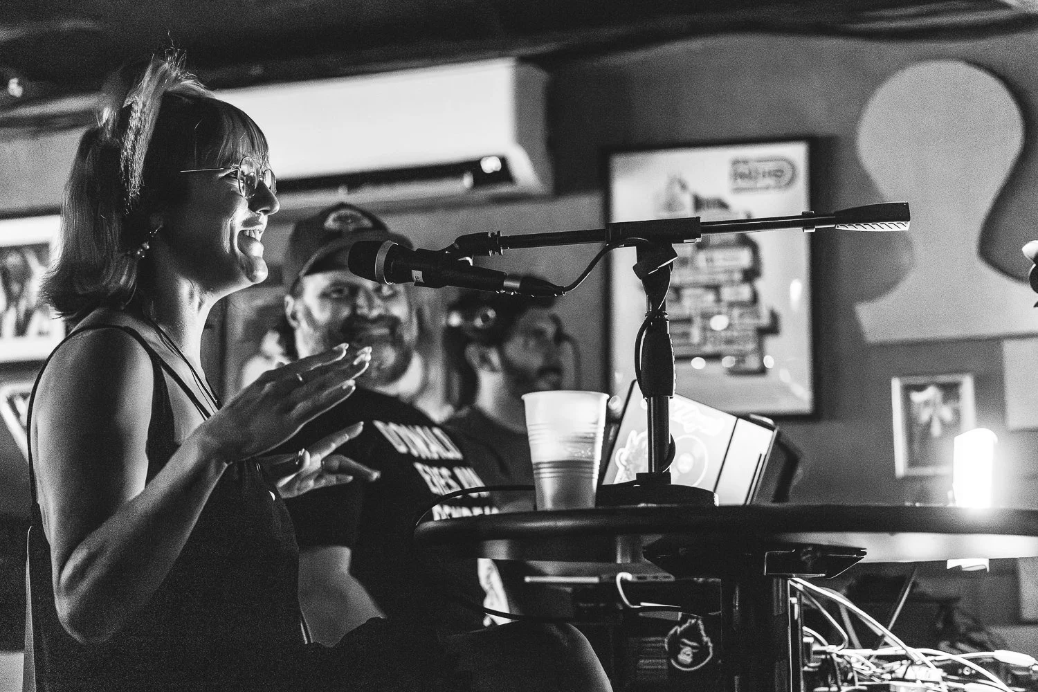 A woman wearing glasses and a sleeveless top is speaking into a microphone at a gathering or event, with two men in the background and various items like cups, a laptop, and papers on the table.