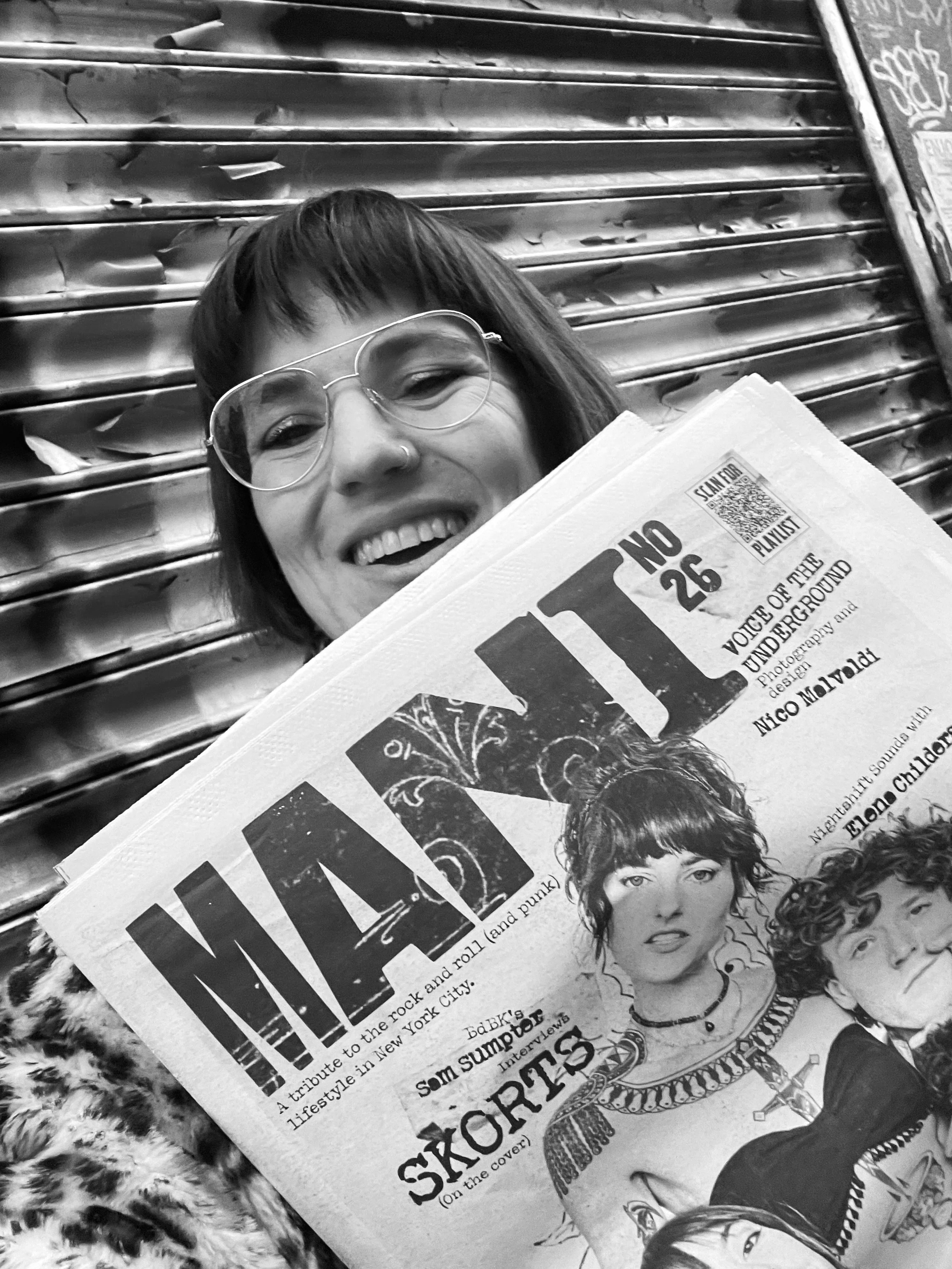 A woman with glasses and short hair smiling while holding a newspaper. She is standing in front of a metal shutter with graffiti.