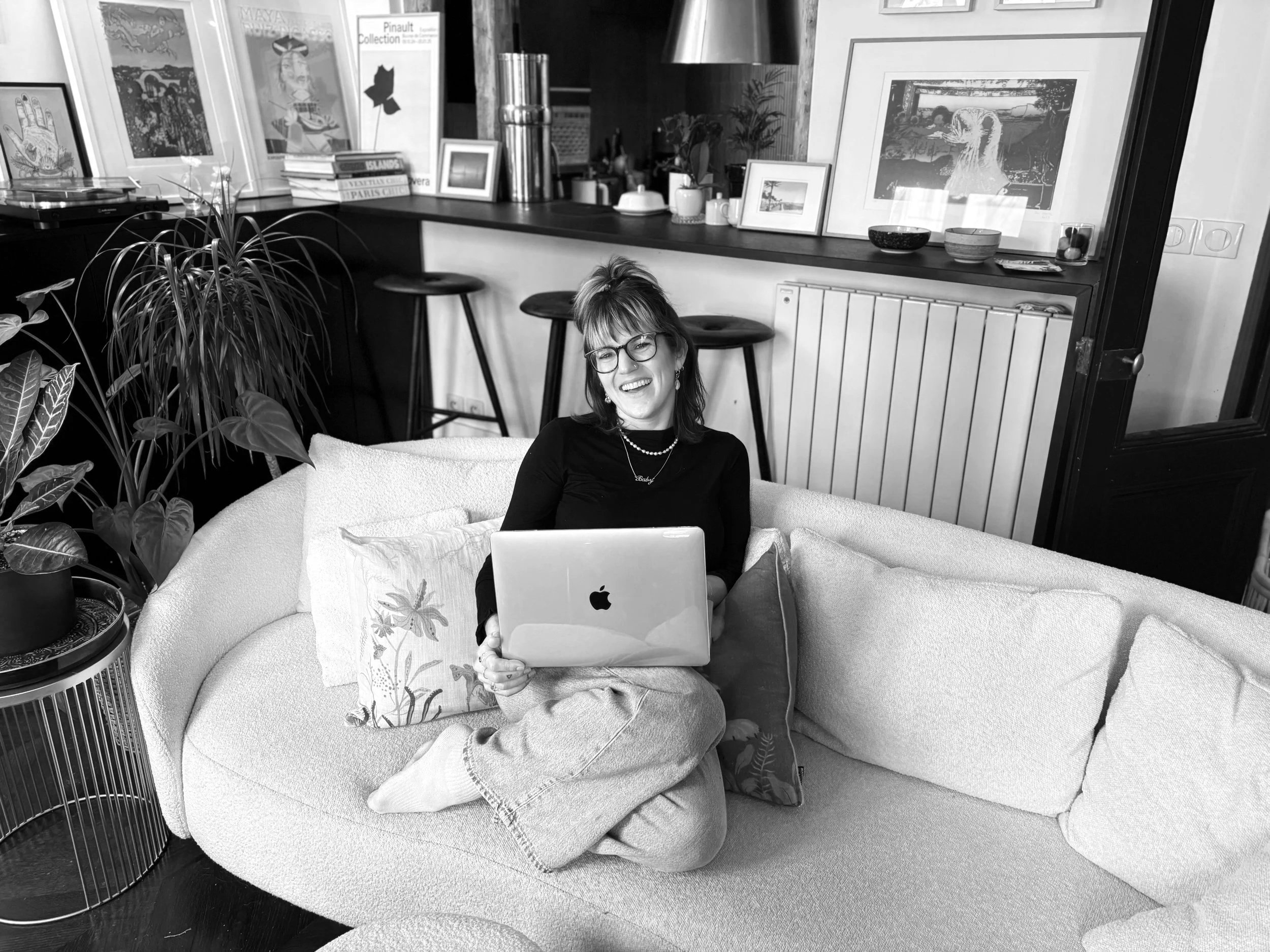 A woman sitting cross-legged on a couch, smiling and holding a laptop, in a living room with plants and framed artwork.