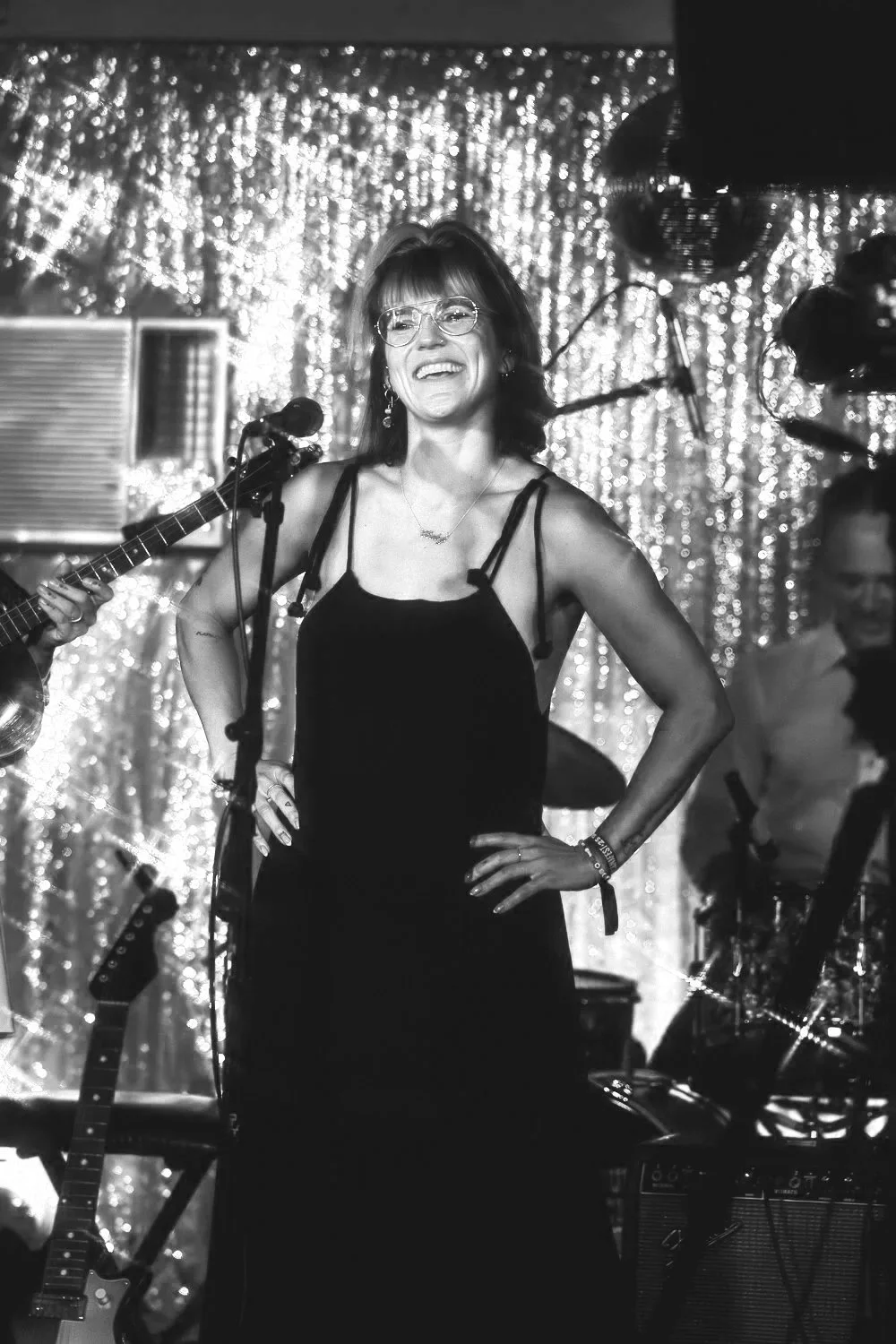 A woman with glasses and a sleeveless black dress standing on stage with a microphone, smiling with hands on her hips, surrounded by musical instruments and performers, with a sparkling backdrop.