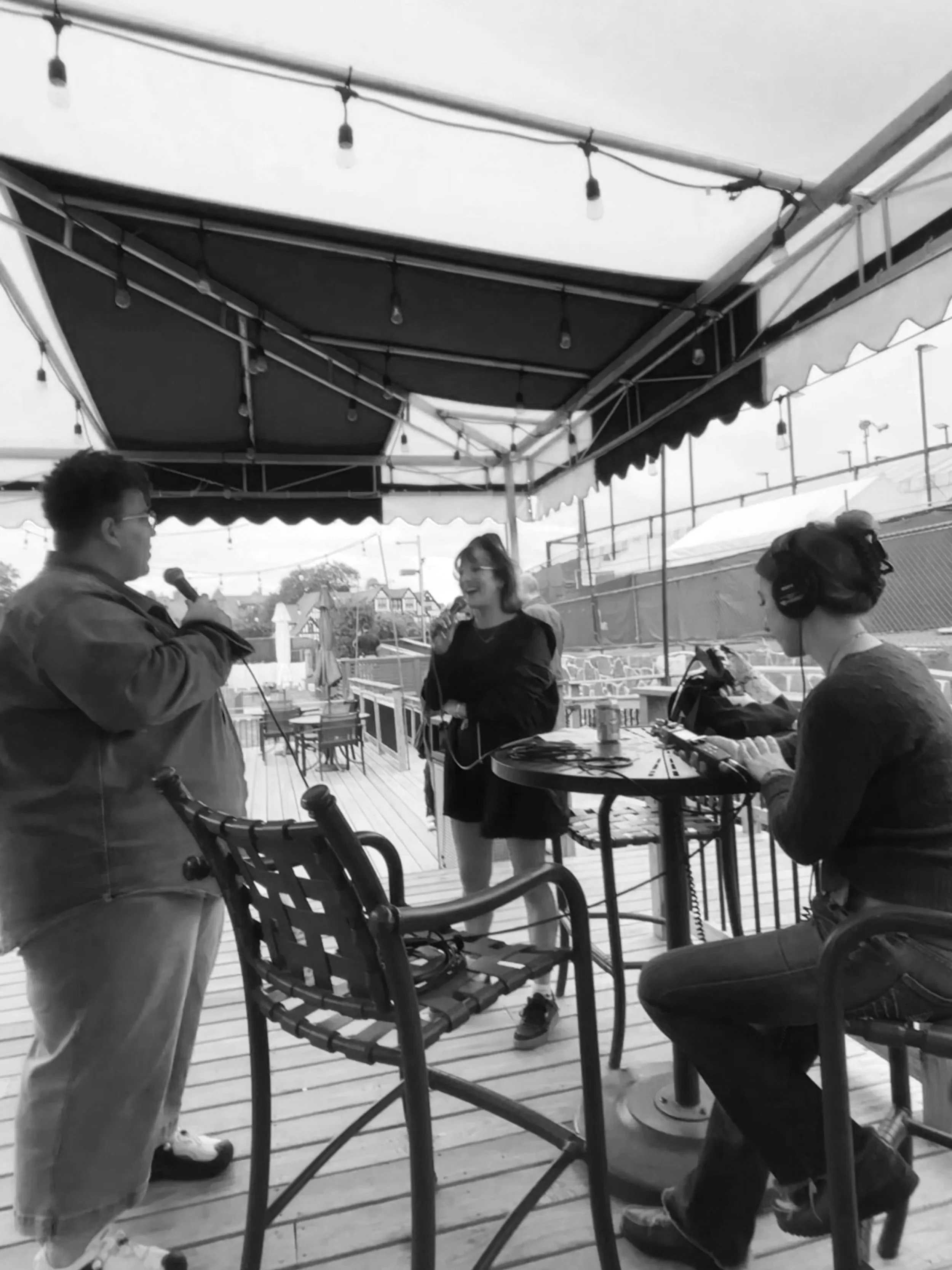 A black and white photo of a woman singing into a microphone, with a man holding a microphone standing nearby, on a covered outdoor patio. A woman wearing headphones operating audio equipment is seated at a round table.
