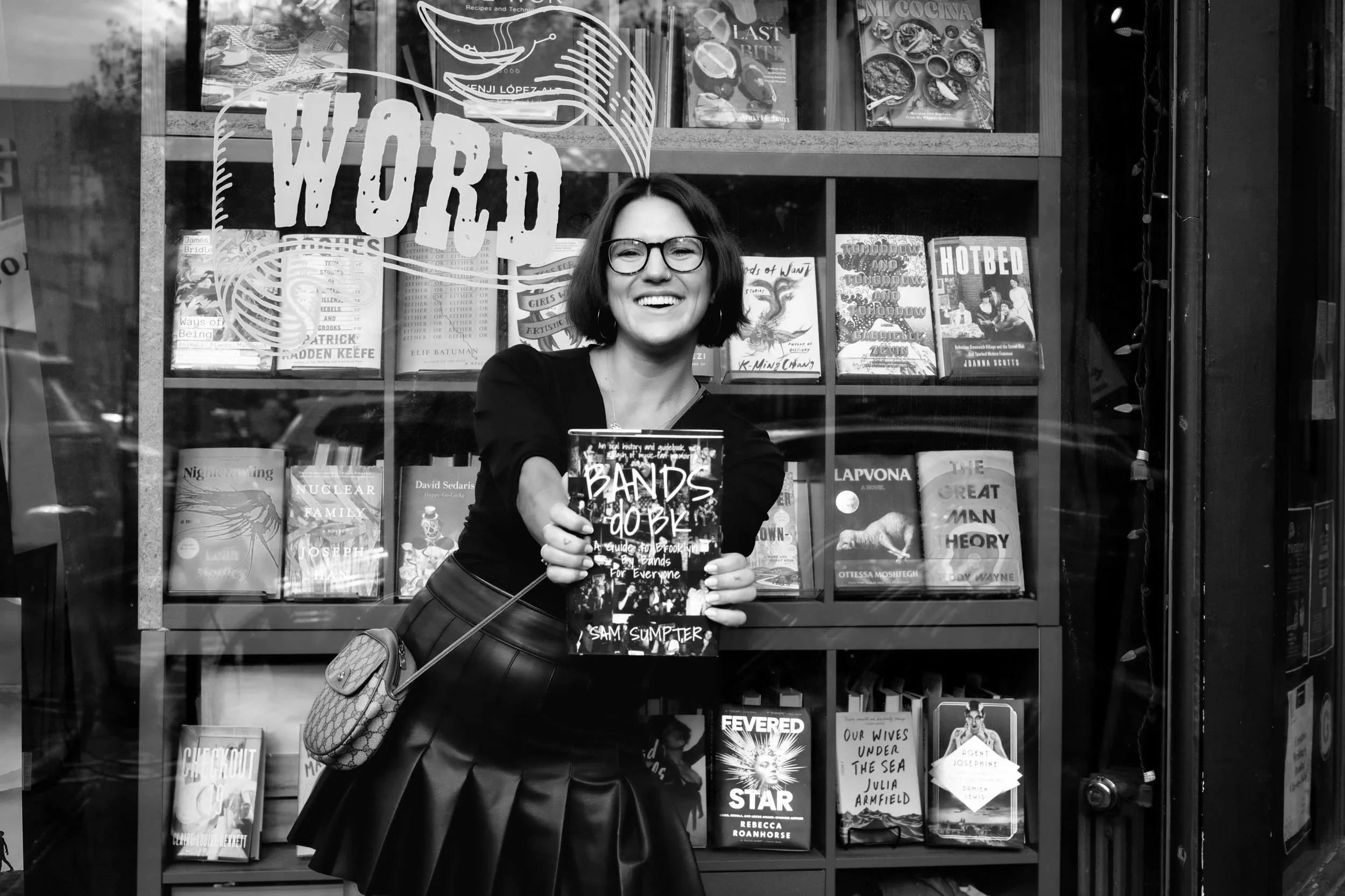 A woman with short dark hair, glasses, wearing a black top and a pleated black skirt, smiling and holding a book titled 'BANDS DO BKE' in front of a bookstore window filled with various books.
