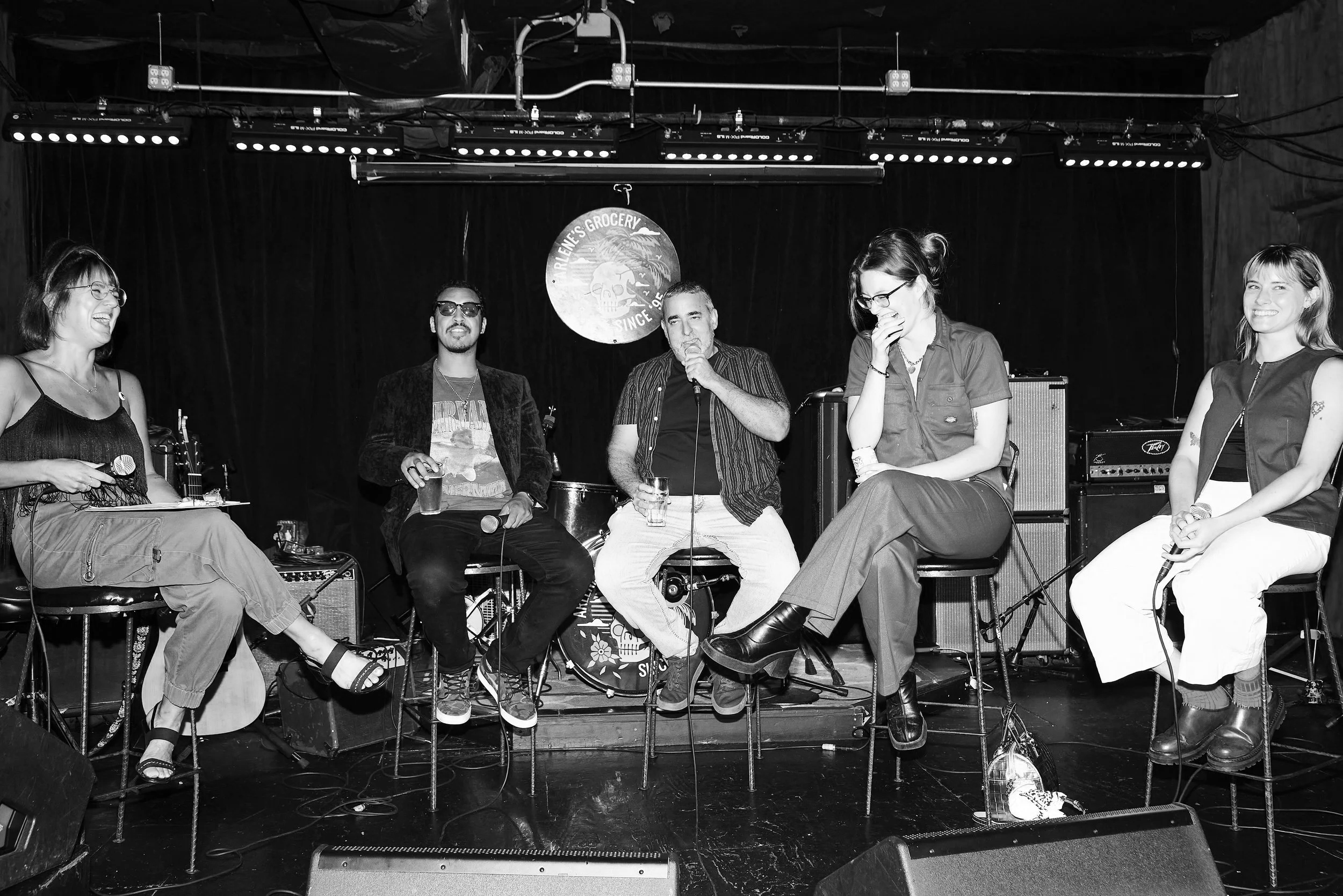 Group of five people sitting on stools on stage during a performance or discussion, with musical equipment and a dark background.
