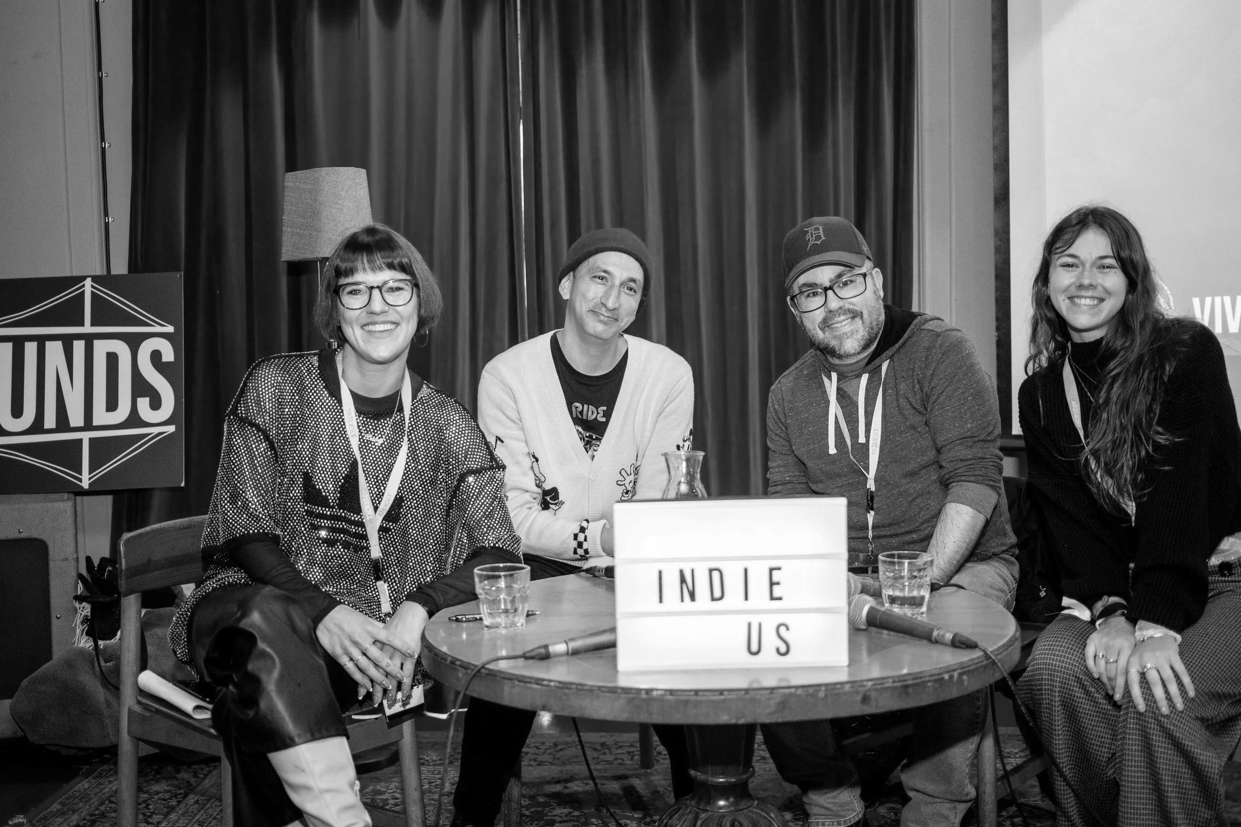 Four people sitting at a round table during an event. They are smiling, with a sign that reads "INDIE US" in front of them. Two women and two men, with microphones and glasses of water on the table.