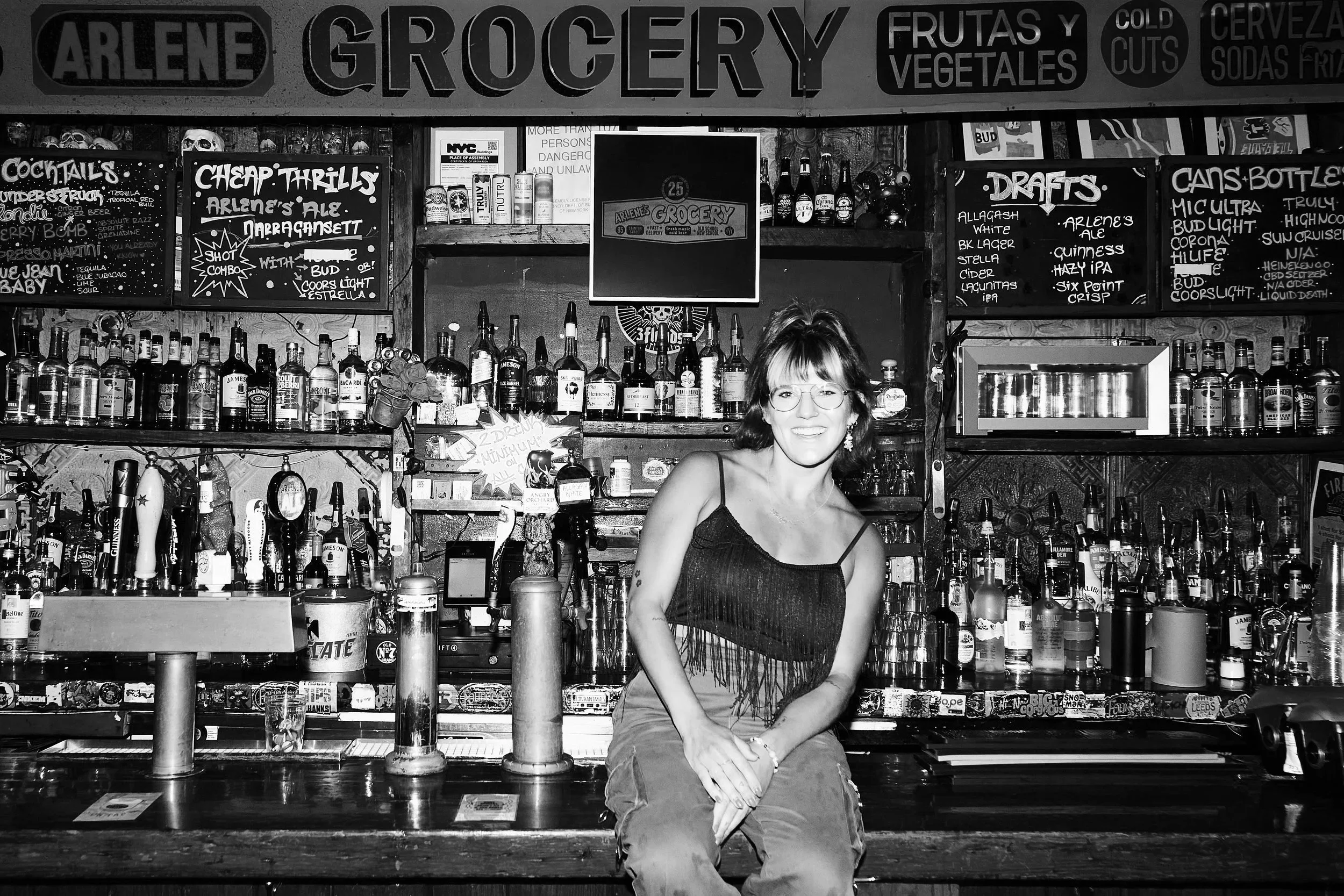 A woman sitting at a bar with bottles of alcohol behind her, various chalkboard menus and signs advertising drinks and specials, in a black and white photo.