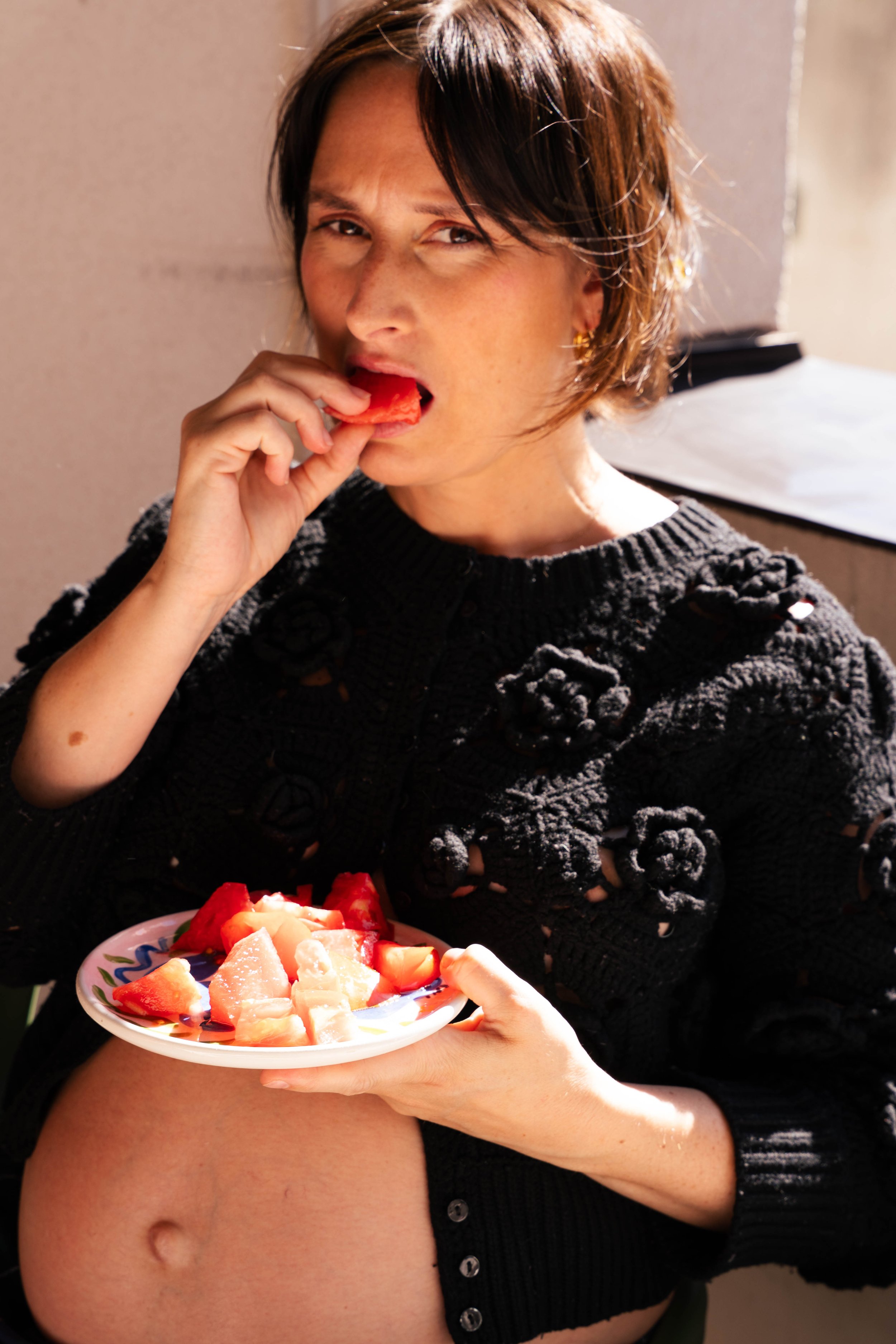 A woman with short brown hair, wearing a black knit sweater, is eating a slice of watermelon. She is holding a plate of cut fruit, including watermelon and pineapple, and is sitting indoors with natural light.