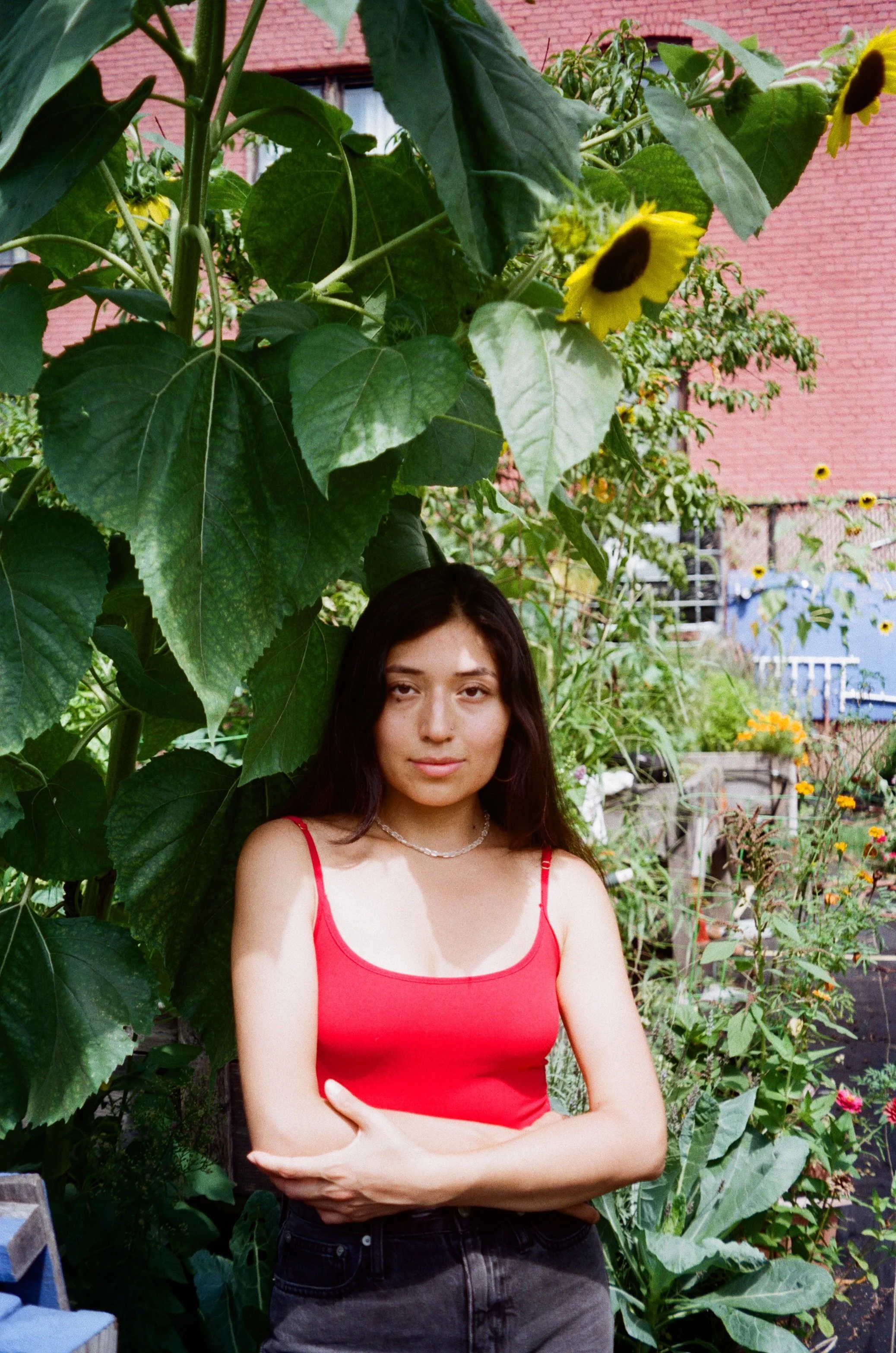 A young woman with long dark hair wearing a red tank top and black pants, standing outdoors in a garden with large green leaves and yellow flowers, a brick building and a red wall in the background.