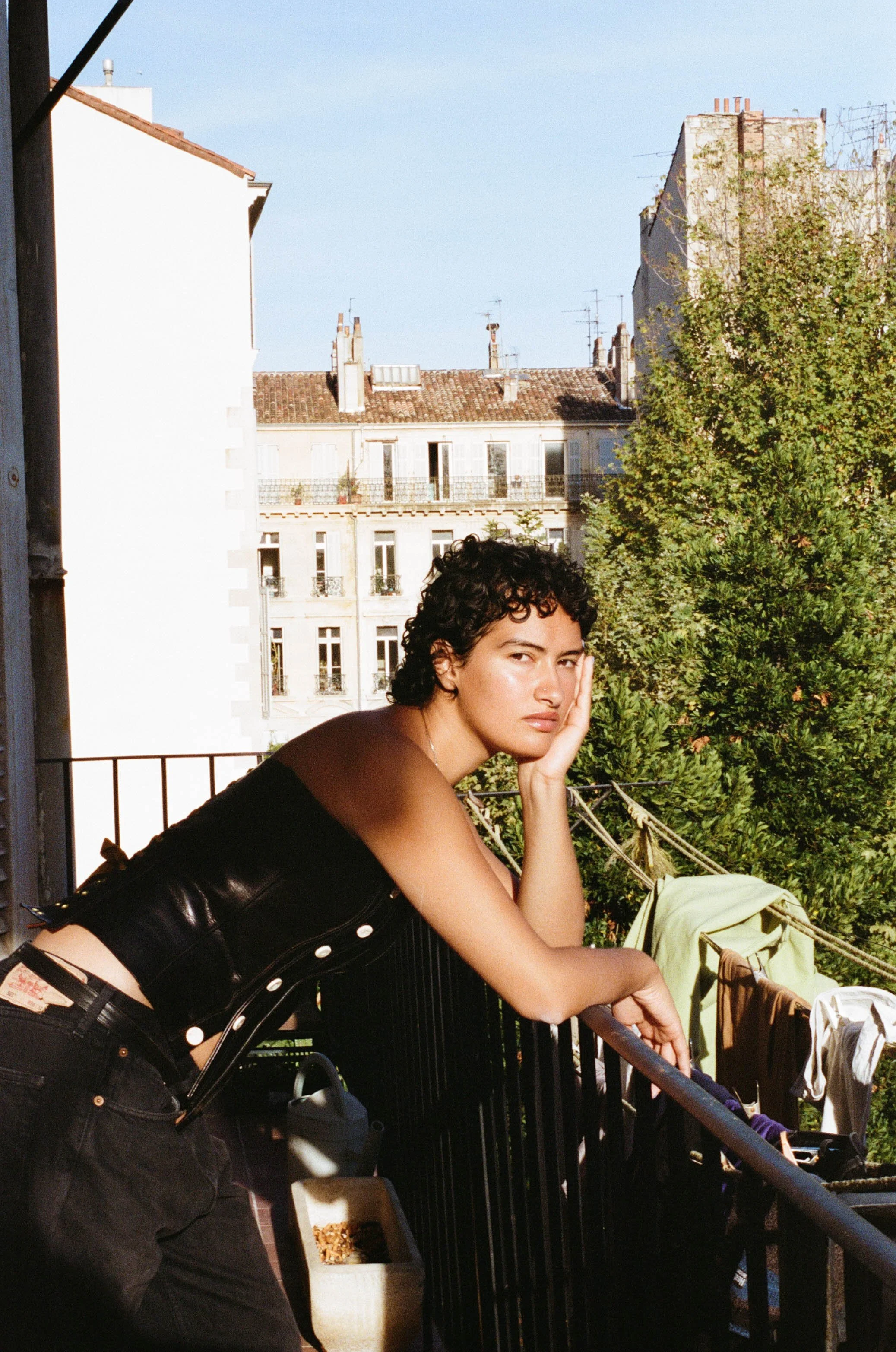 A young woman with curly dark hair and light skin is leaning on a balcony railing, resting her face on her hand. She is wearing a black cropped top and black pants, with a belt. The balcony has laundry hanging on a line, and there are trees and multi-story buildings with rooftops and chimneys in the background.
