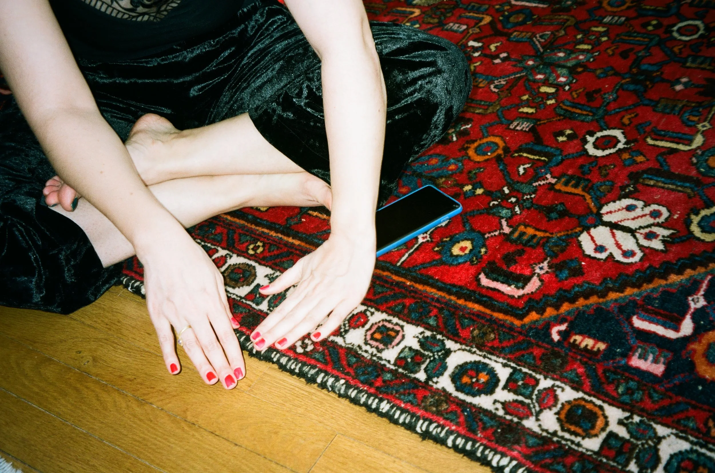 A person with pale skin is sitting cross-legged on a red patterned rug on a wooden floor. They are wearing black velvet pants and a black top, with their hands resting on the floor, showing red nail polish. A smartphone is placed on the rug near their hand.
