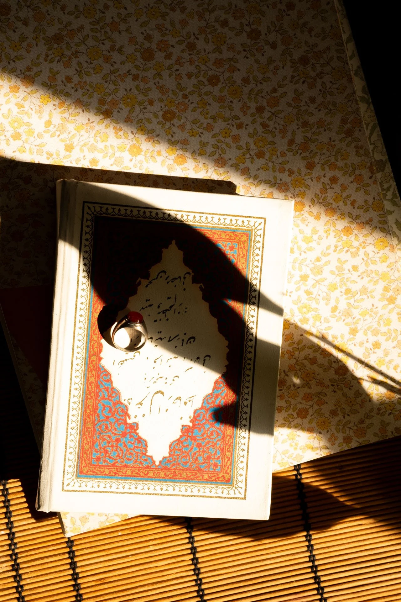 A paperback book with ornate border and Arabic script, a silver ring with a red gemstone on top, and a shadow cast over the book, all placed on a floral patterned cloth and bamboo mat.