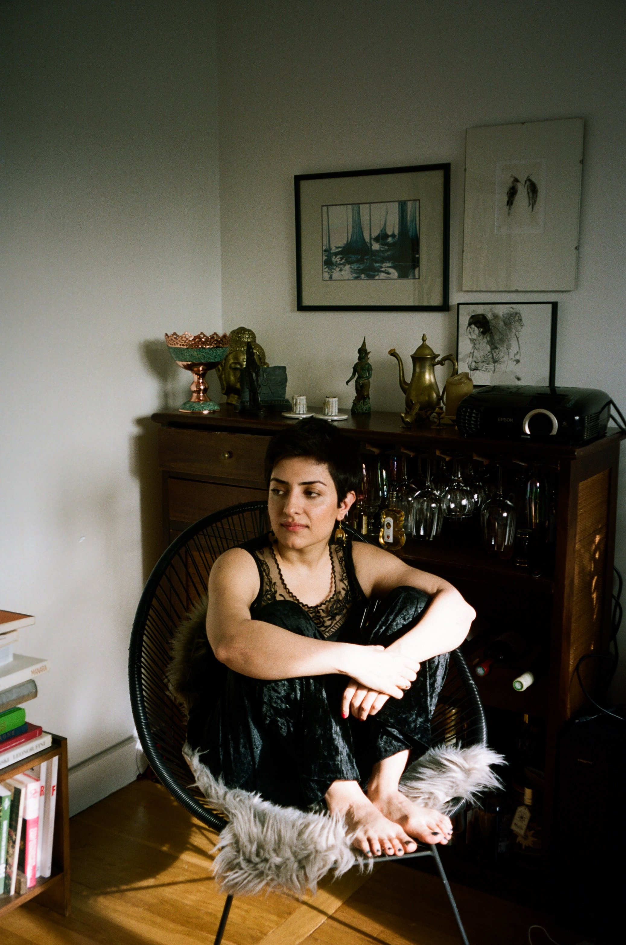 A woman with short dark hair sitting in a modern black wire chair with a faux fur throw on the seat, cross-legged, wearing a black lace top and velvet pants, in a cozy room with a wooden sideboard and framed art on the wall, and various decorative objects including sculptures and a brass teapot visible behind her.