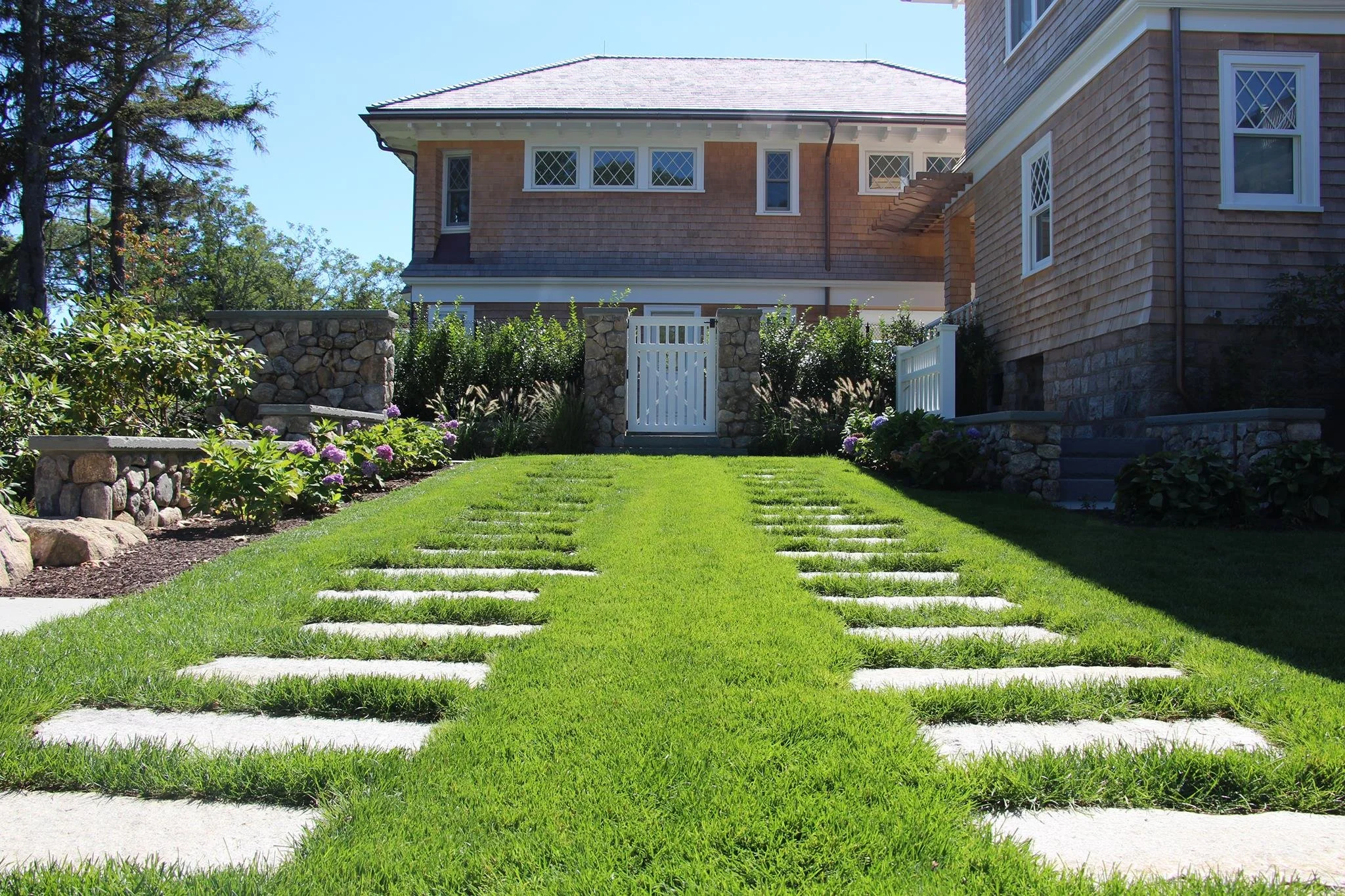 A well-maintained front yard with a green grass pathway of rectangular stone slabs leading to a white gate, flanked by flower beds and stone walls, with a house in the background.