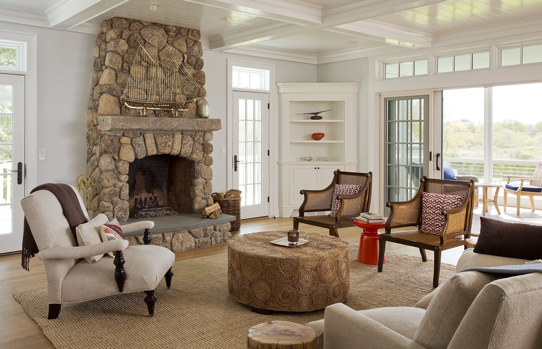 Living room with a stone fireplace, beige armchairs, wooden chairs, a round ottoman, and large windows with views of a patio and greenery.
