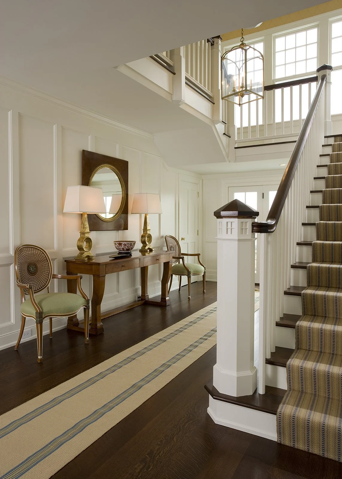 Entryway of a house with white paneled walls, a dark hardwood floor, a beige striped runner rug, a wooden console table with lamps and a decorative bowl, French style chairs, a staircase with dark wood steps and a patterned runner, and a second floor balcony with white railing and large windows.