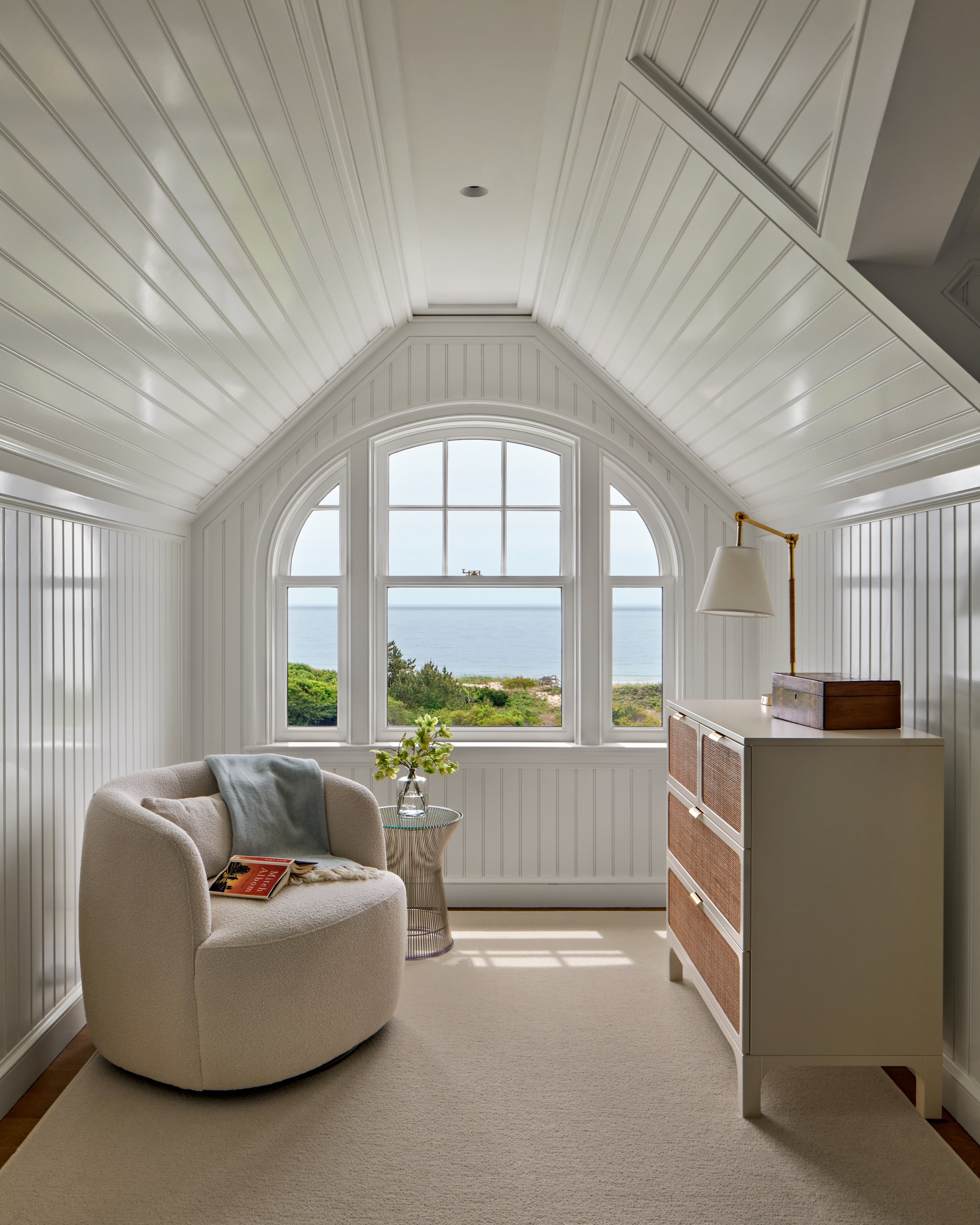A small cozy room with a large arched window overlooking the ocean, white paneling on the walls and ceiling, a round beige armchair with a light blue throw blanket and a magazine, a small side table with a glass vase and green plant, and a white dres