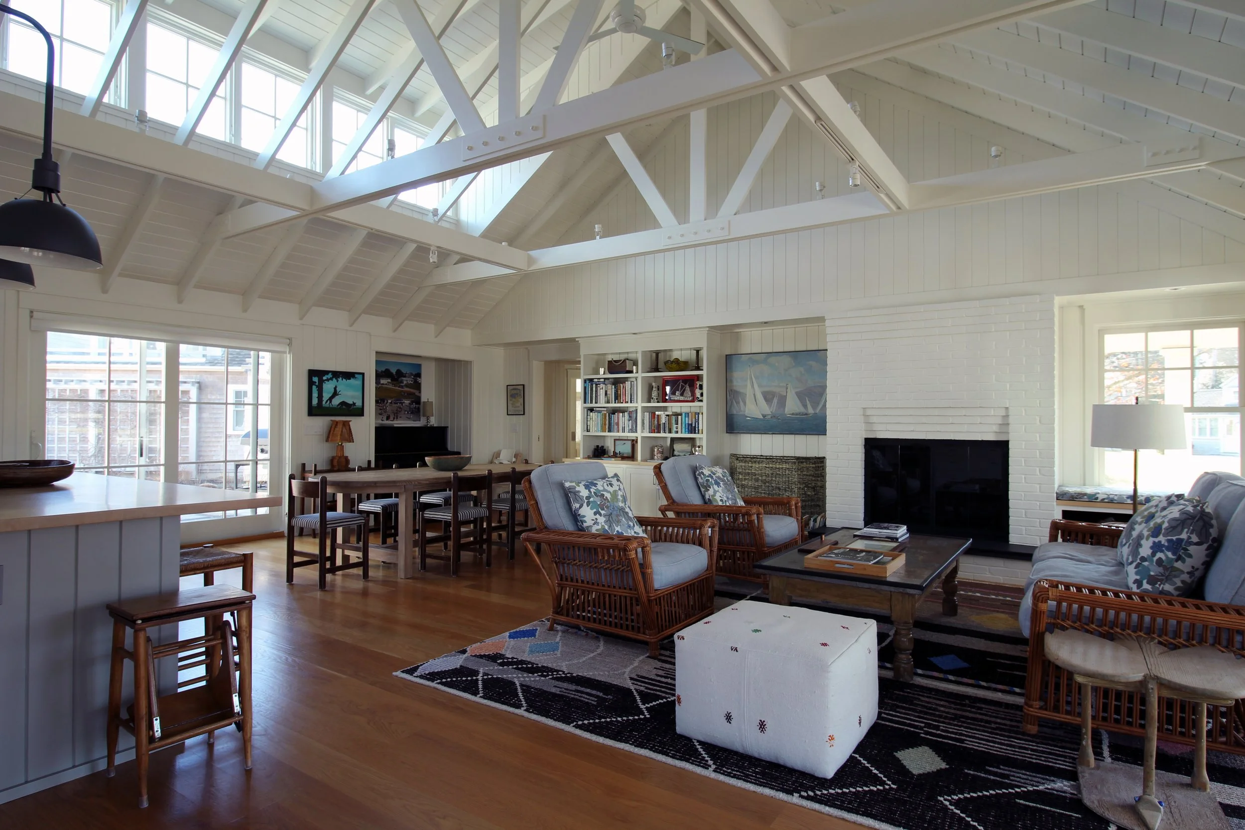 Bright living room with white vaulted ceiling, skylights, white brick fireplace, wooden furniture, patterned area rug, and large windows.