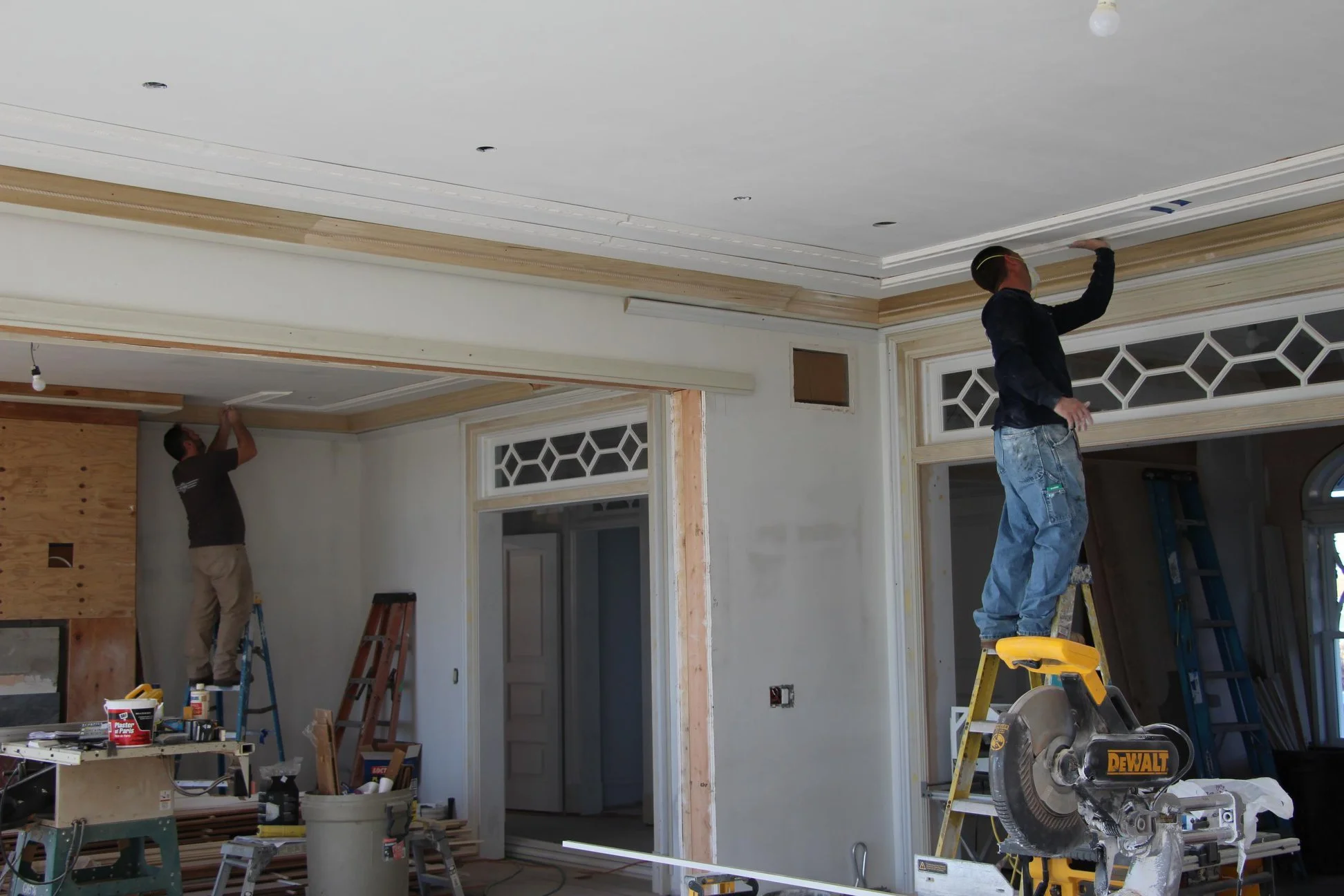 Two workers renovate a room, one standing on a yellow ladder and the other on a chair, installing or repairing the ceiling.