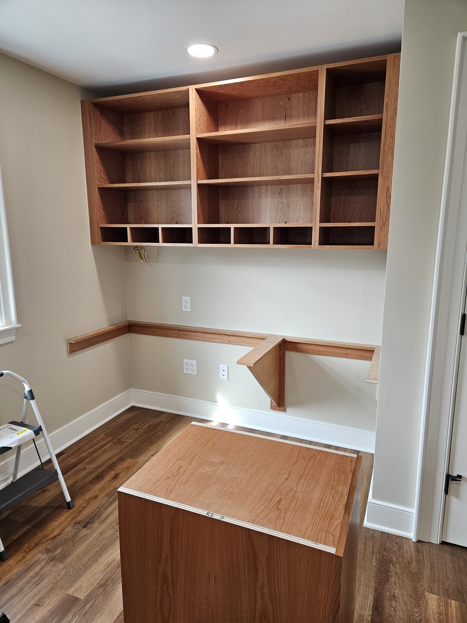 unfinished built-in wooden shelves and desk in a room with hardwood floors and beige walls.