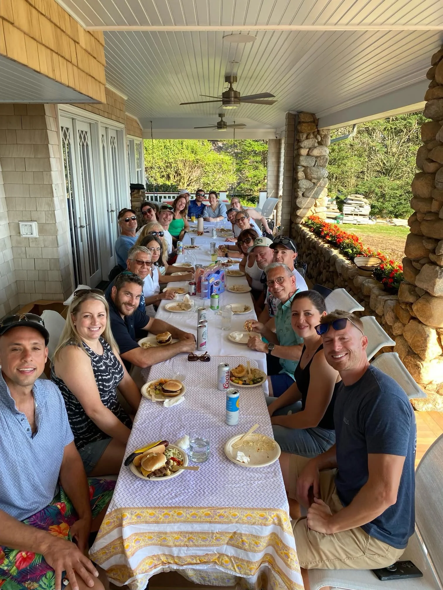 A large group of people gathered around a long outdoor dining table on a covered porch, enjoying a meal together.