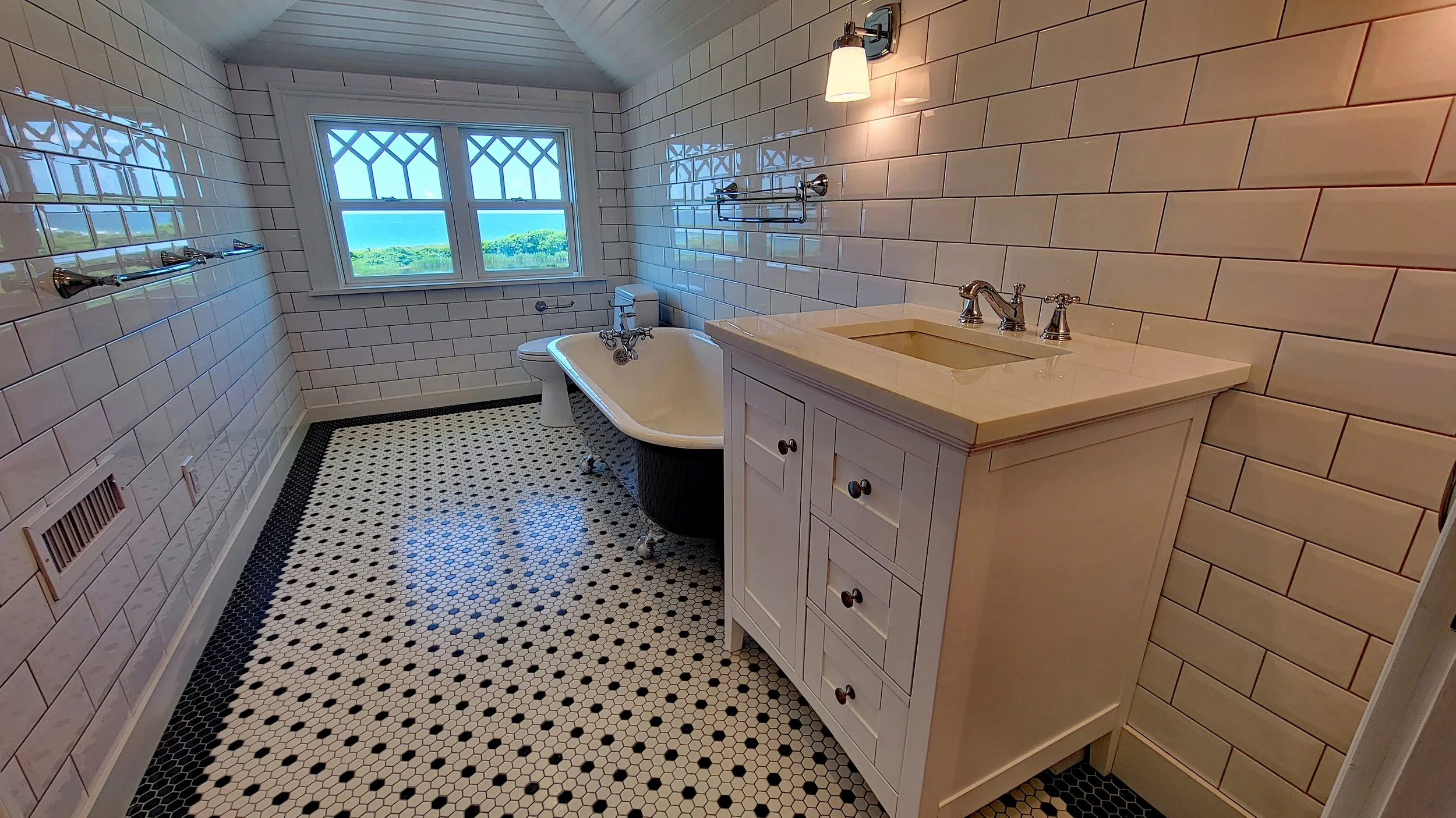 A vintage bathroom with white tiled walls, black and white hexagonal floor tiles, a clawfoot bathtub, a white vanity with a sink, a window with a scenic view, and modern light fixtures.