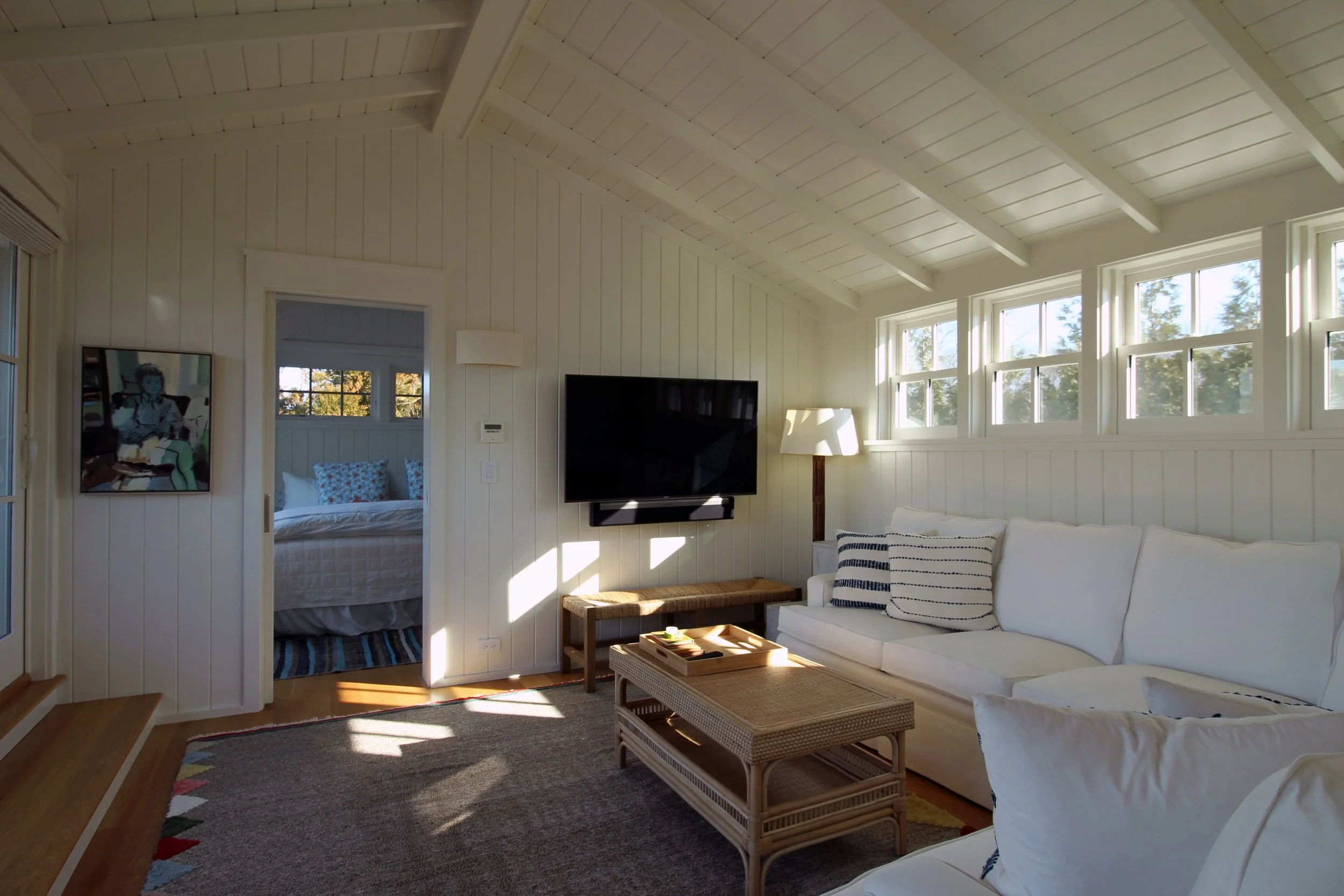 Bright living room with white walls and ceiling, large windows, white sofa with striped pillows, coffee table, wall-mounted flat-screen TV, standing lamp, view into a bedroom with bed and pillows.