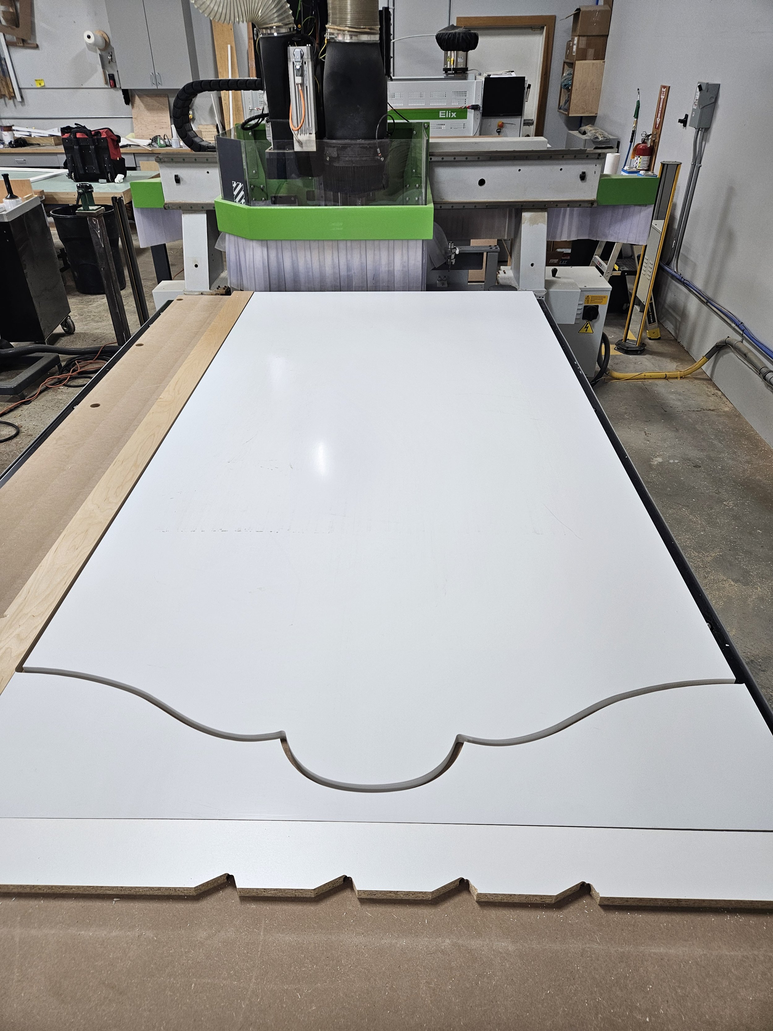 A large sheet of white material with a decorative cutout edge on a worktable in a workshop. A CNC or laser cutting machine is positioned at the back of the worktable, with hoses and mechanical components visible.