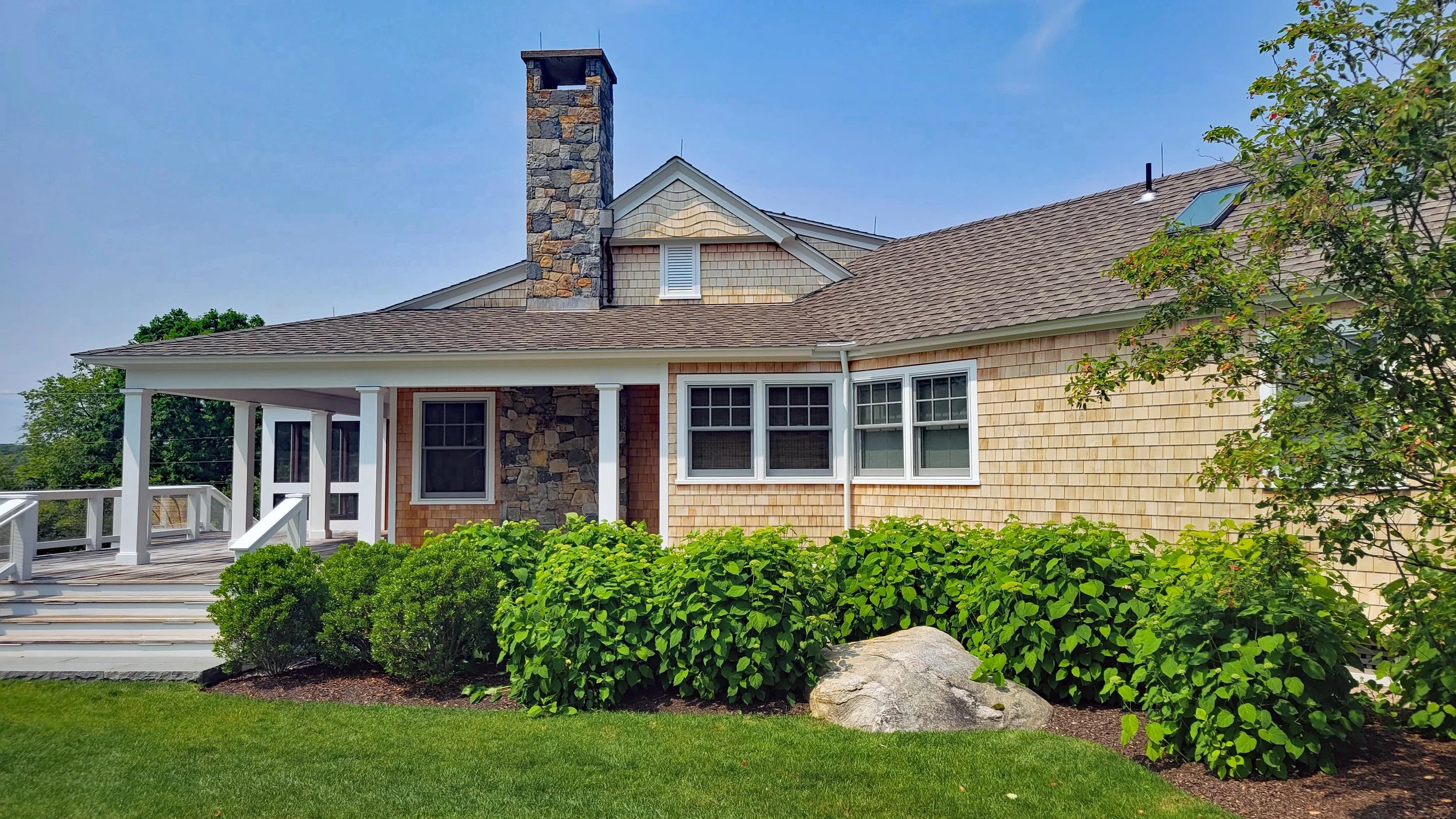 Exterior of a house with a stone chimney, shingle siding, and a deck with white railings, surrounded by shrubs and greenery under a clear blue sky.