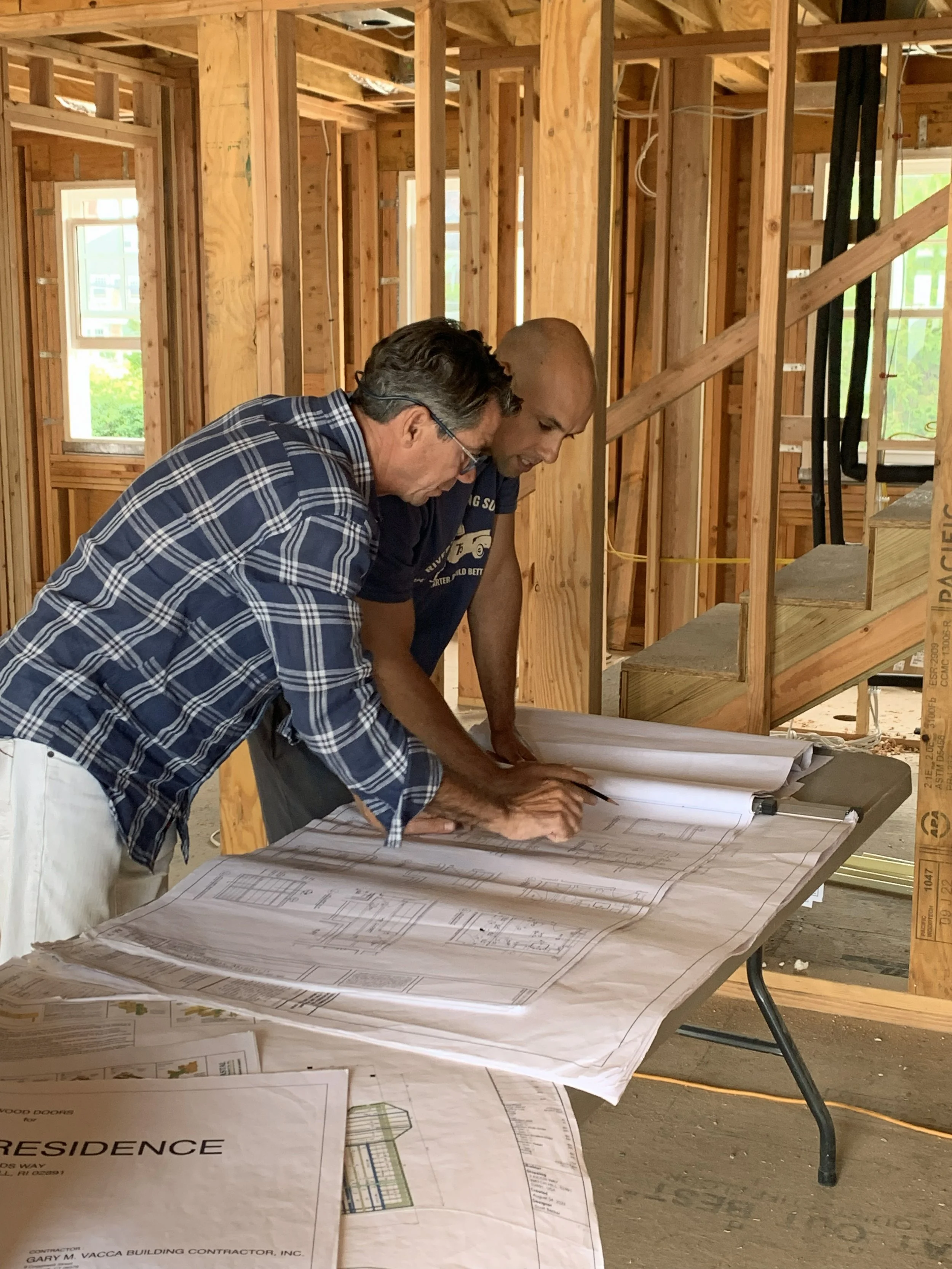 Two men examining and discussing architectural blueprints on a table inside a partially constructed house with exposed wooden framing.