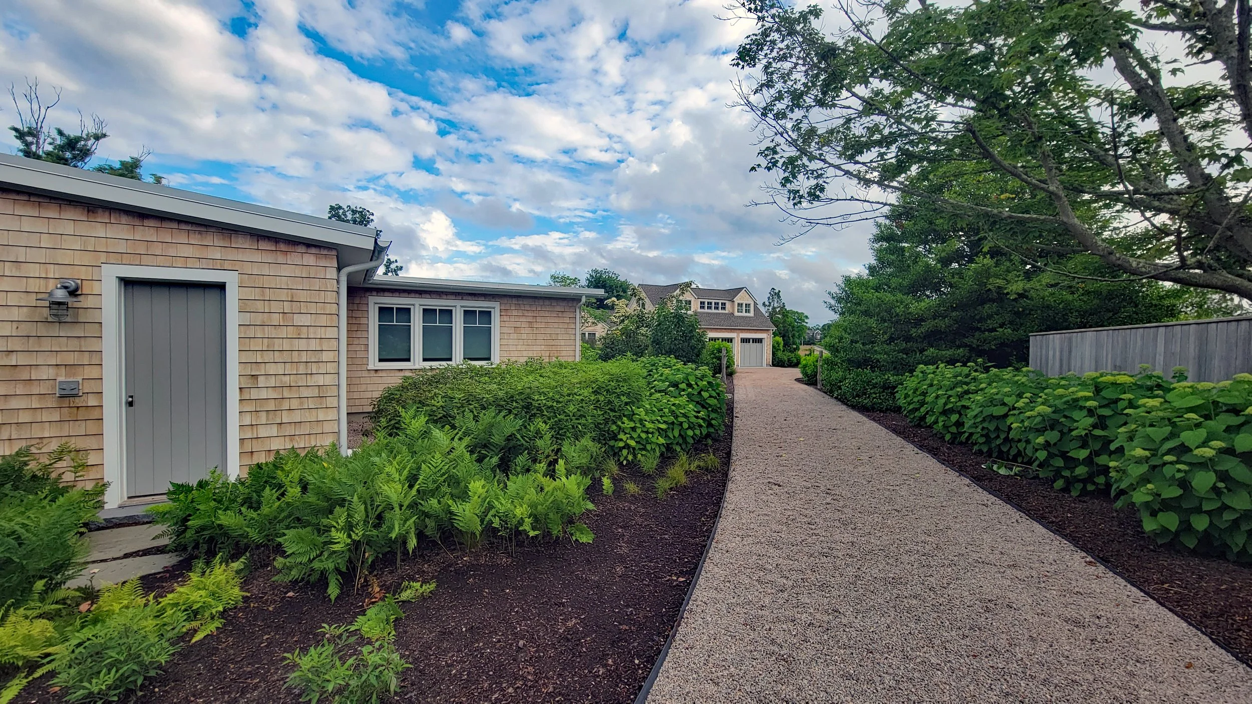 A gravel pathway leading to a garage, lined with green shrubs and trees, under a partly cloudy sky.