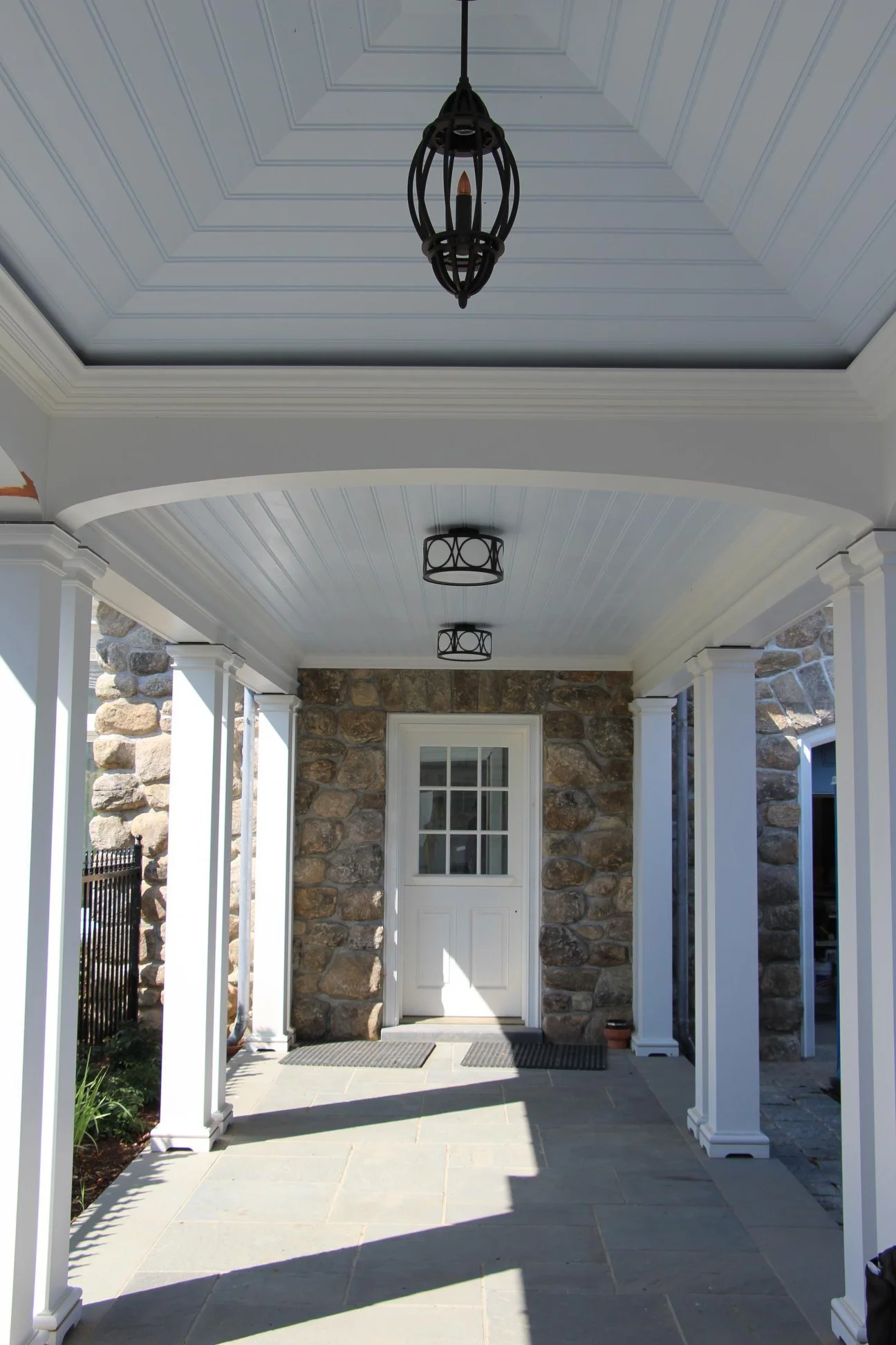 Covered porch with stone wall, white columns, and ceiling lights, leading to a door with a window.