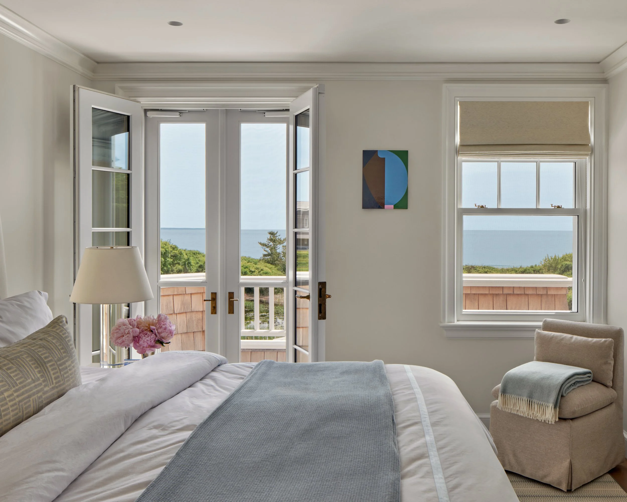 A cozy bedroom with white walls and large windows overlooking a scenic view of water and greenery, featuring a bed with white linens, a gray blanket, a lamp, a small table with pink flowers, and a beige chair with a rolled blanket.