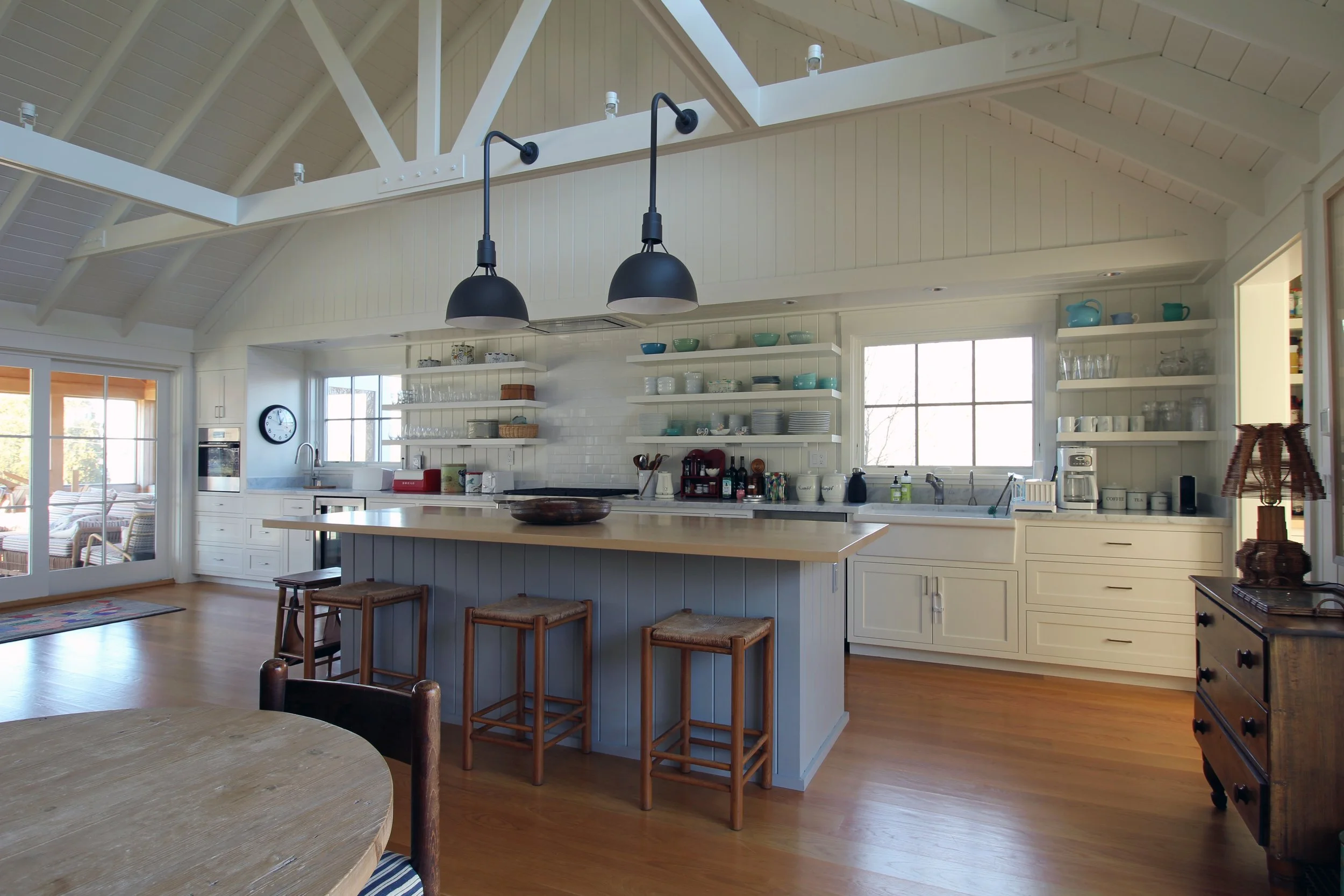Bright kitchen with white cabinets, open shelves with dishes, wooden island with stools, hardwood floors, large window, ceiling beams, and sliding glass door.