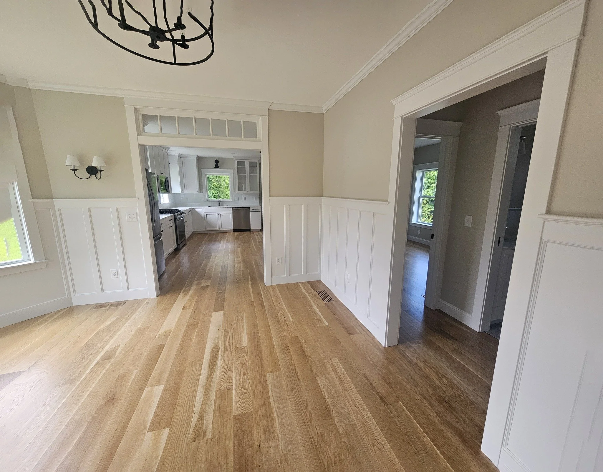 Empty dining room and kitchen with hardwood floors, white wainscoting, and large windows.