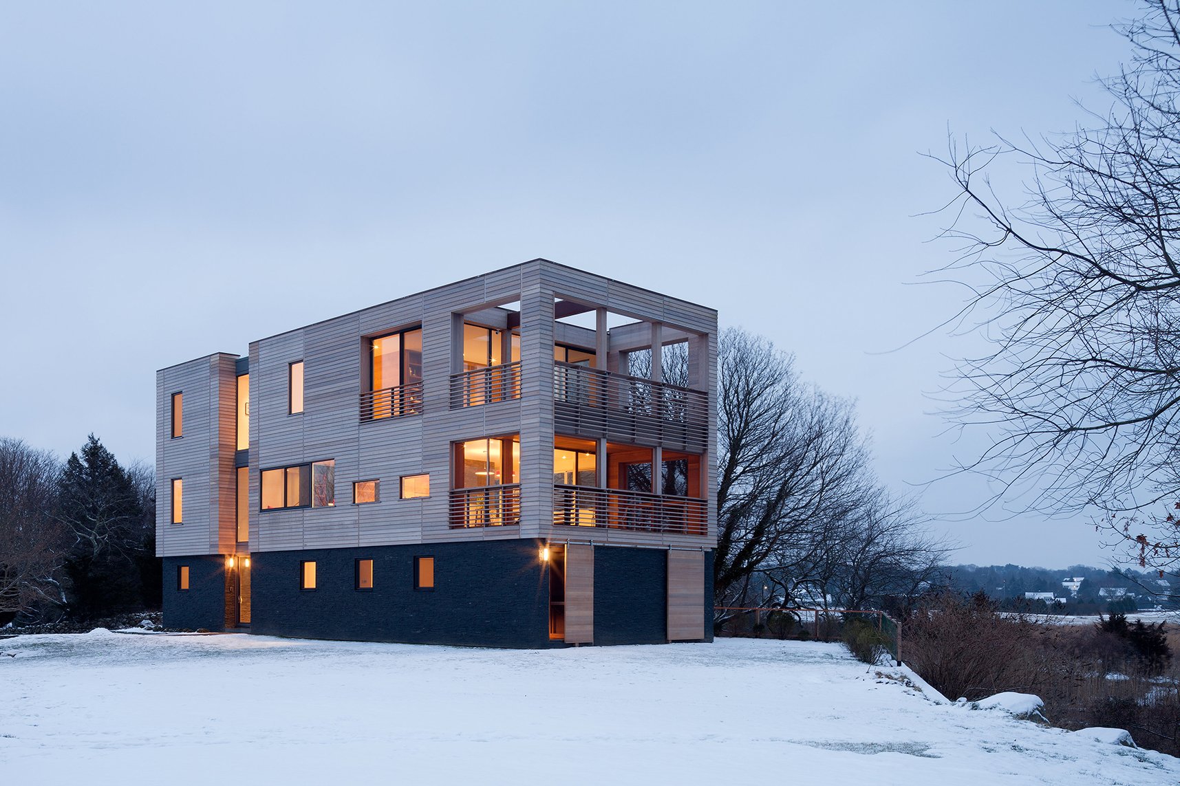 Modern three-story house with large windows and balconies, surrounded by snow and leafless trees during winter evening.