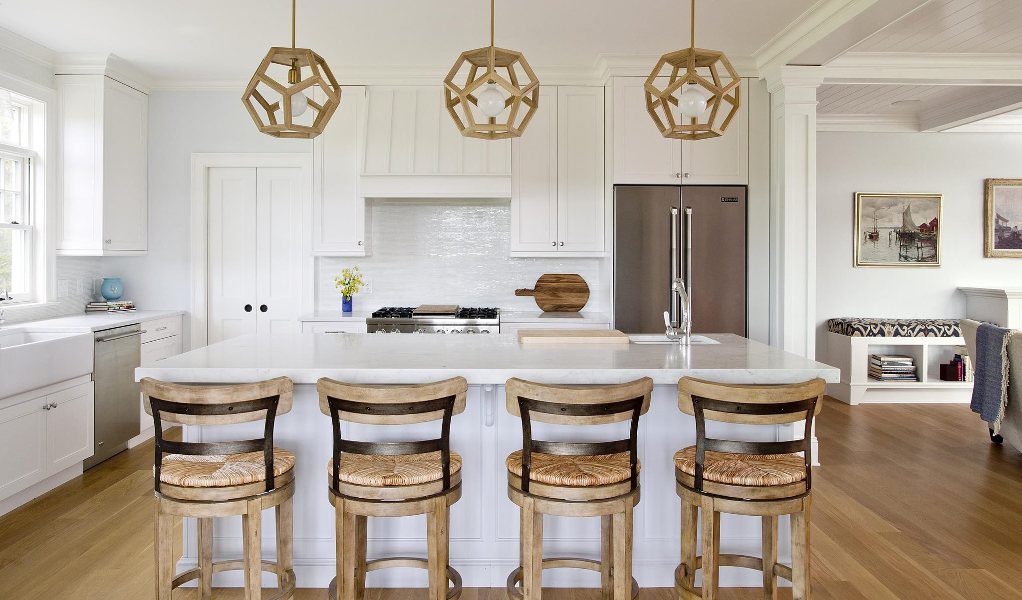 Modern kitchen with white cabinets, a marble island, wooden bar stools, pendant lights, a stainless steel refrigerator, and artwork on the walls.